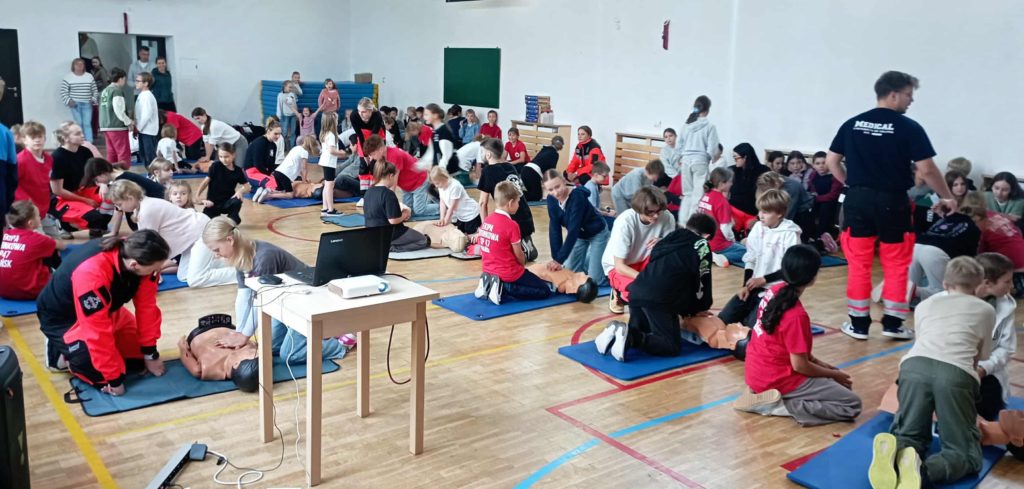 W sali gimnastycznej na podłodze leżą 3 rzędy fantomów przy nich klęczą po 2 lub 3 osoby. Jedna z osób wykonuje na fantomie masaż serca. Na pierwszym planie stolik z laptopem i rzutnikiem. W tle wyjście z sali.