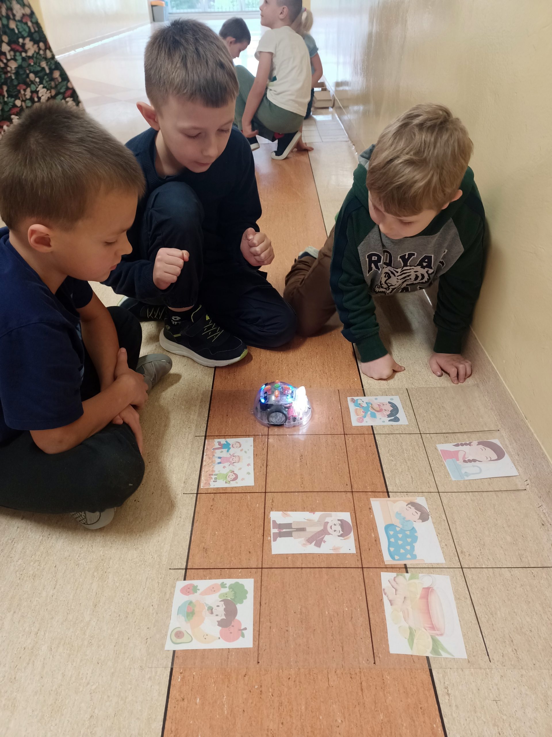 3 uczniów 1a siedzi na szkolnym korytarzu przy planszy z obrazkami i małym robotem edukacyjnym do kodowania.