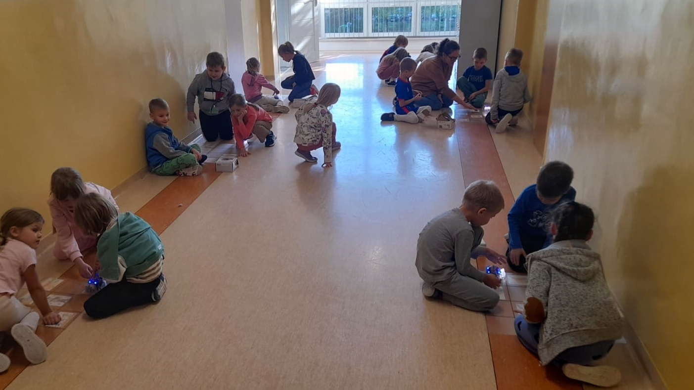 Grupa dzieci z 1b pracuje w parach i małych grupach na szkolnym korytarzu, programując roboty edukacyjne na kartach z obrazkami.
