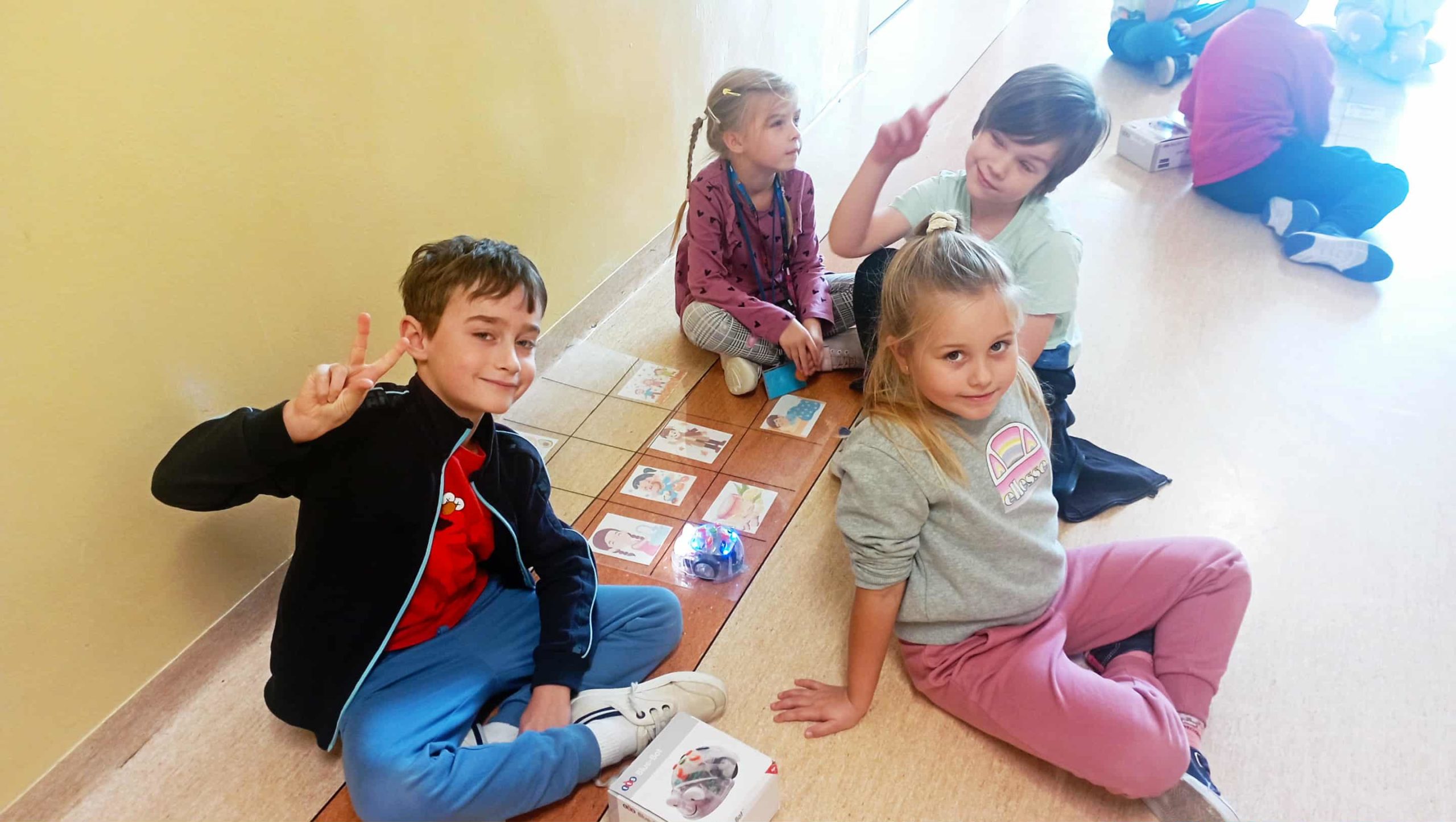 4 dzieci z 1c 3 uczniów 1c siedzi na szkolnym korytarzu przy planszy z obrazkami i małym robotem edukacyjnym do kodowania.