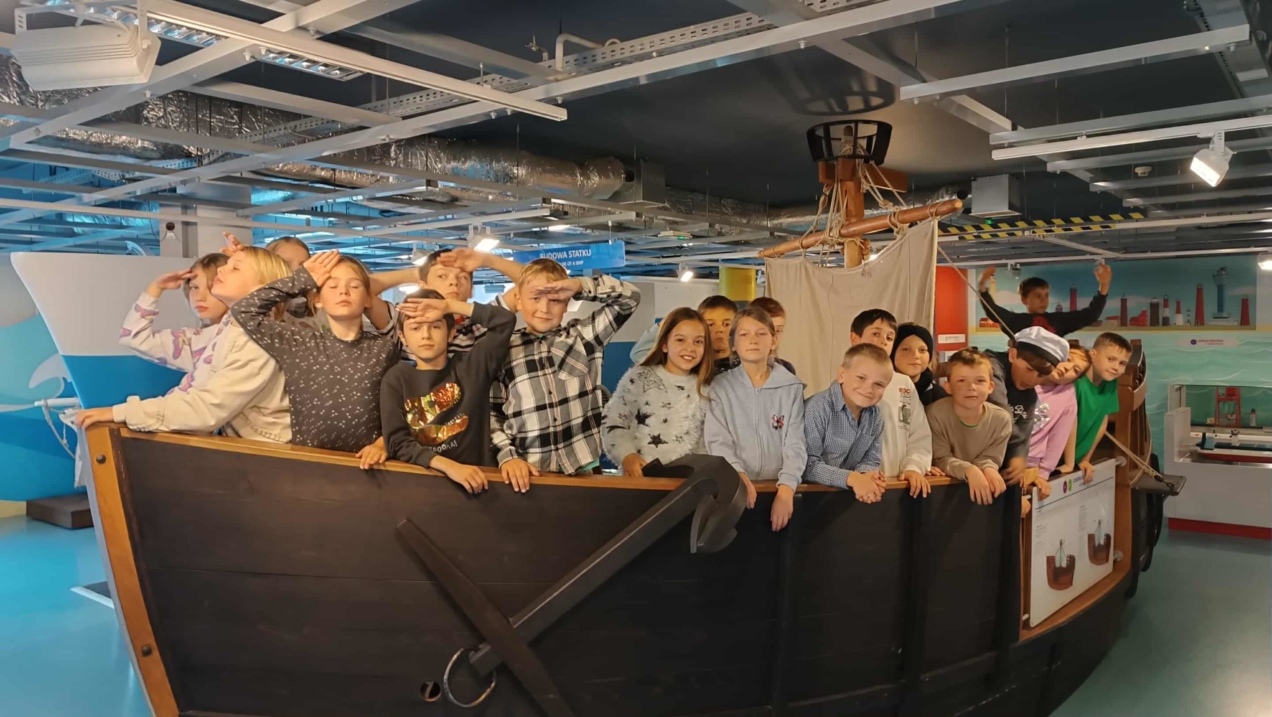 Grupa dzieci stoi na makiecie drewnianego statku w muzeum, część z nich zasalutowała i patrzy w dal, symulując marynarzy podczas rejsu.