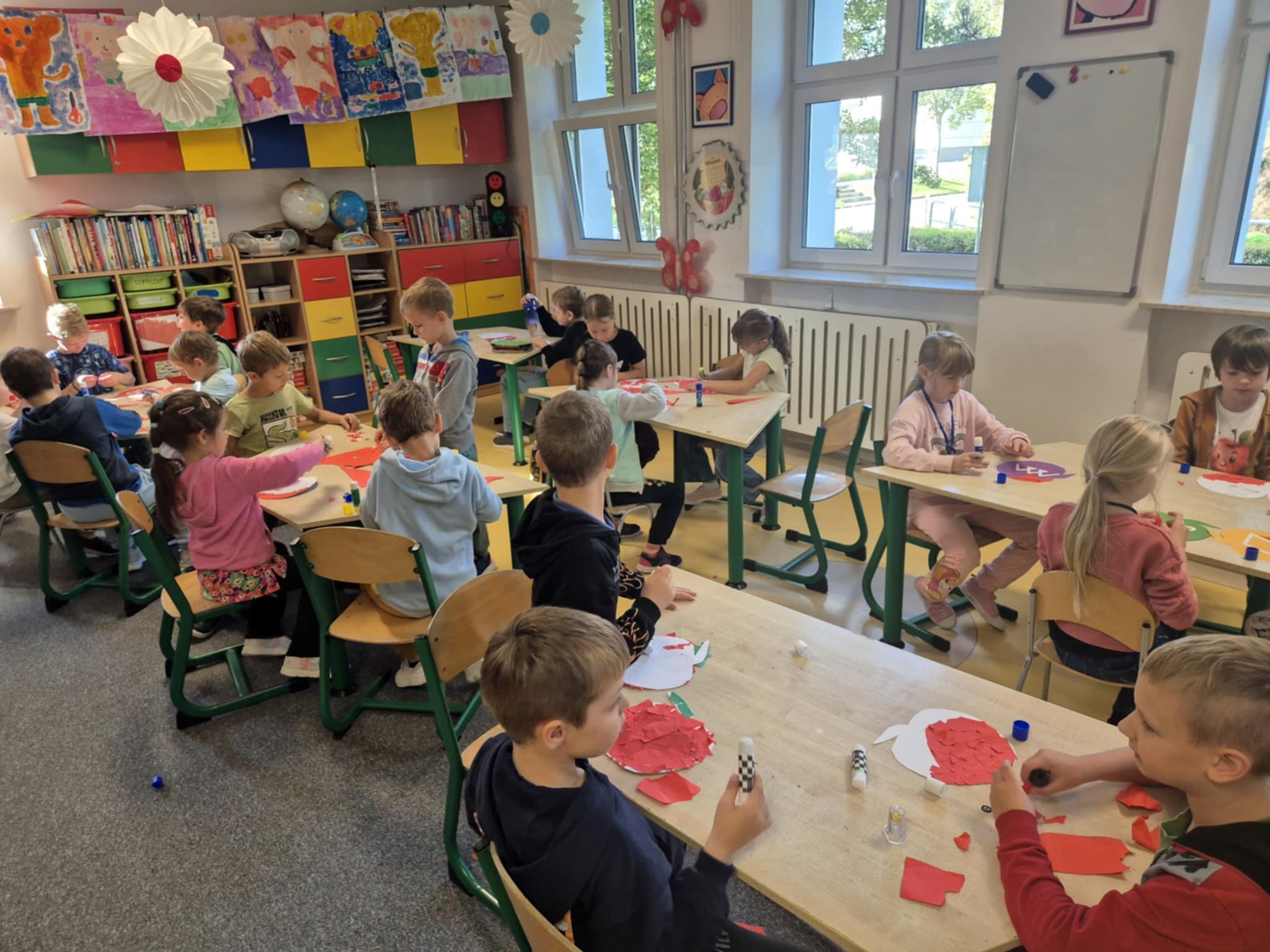 Grupa dzieci siedzi przy szkolnych stolikach i wykonuje prace plastyczne z papieru w kształcie jabłek. W tle kolorowe szafki i zawieszone na sznurku prace plastyczne.