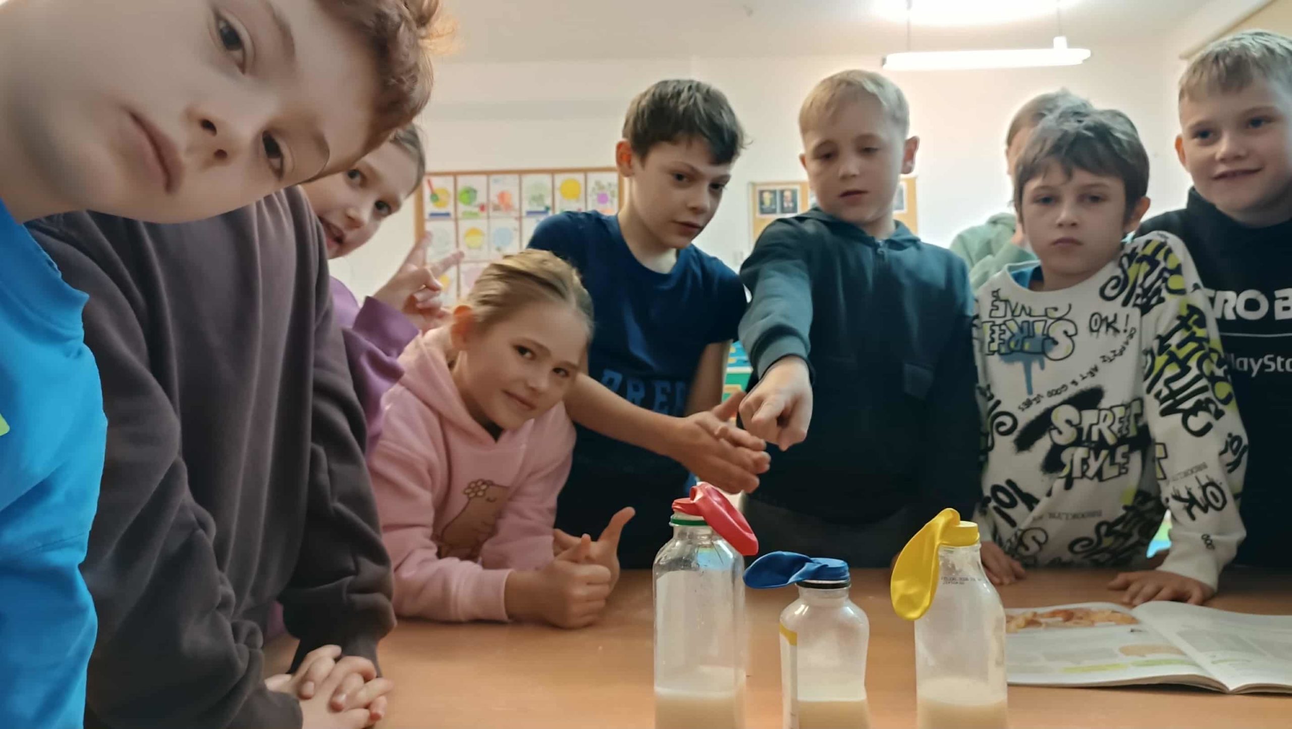 Uczniowie w klasie  z zainteresowaniem obserwują eksperyment z drożdżami – baloniki na butelkach pokazują, jak drożdże produkują gaz.