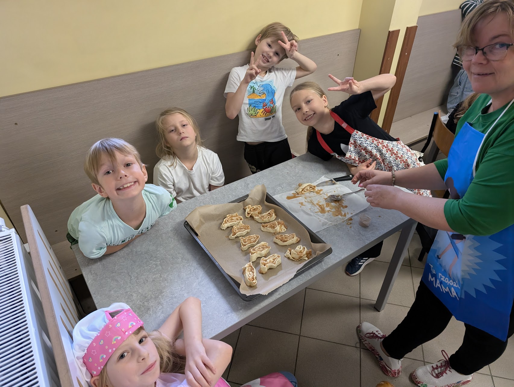 Grupa dzieci i kobieta przygotowująca wypieki w kuchni lub sali warsztatowej. Na stole leżą surowe ciasteczka lub bułeczki ułożone na blasze wyłożonej papierem do pieczenia. Kobieta w okularach i niebieskim fartuchu rozsmarowuje farsz. Dzieci w różnym wieku uśmiechają się i pozują do zdjęcia – niektóre pokazują znak pokoju, inne patrzą z zaciekawieniem. W tle widać ławkę i ścianę w jasnych kolorach.