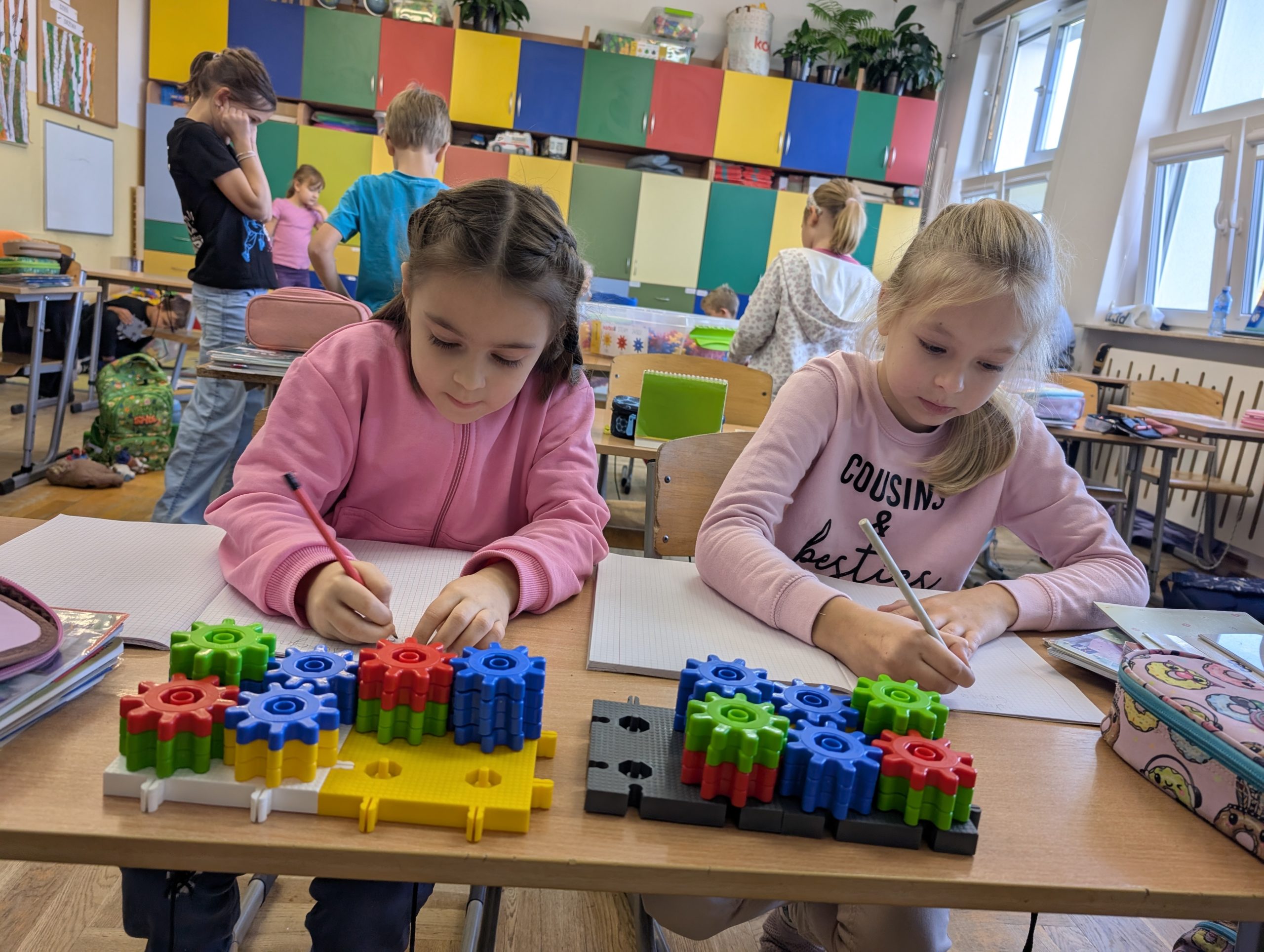 Dwie dziewczynki siedzą przy ławce i zapisują obserwacje w zeszytach. Na biurku leżą kolorowe konstrukcje z klocków Korbo w kształcie trybików.