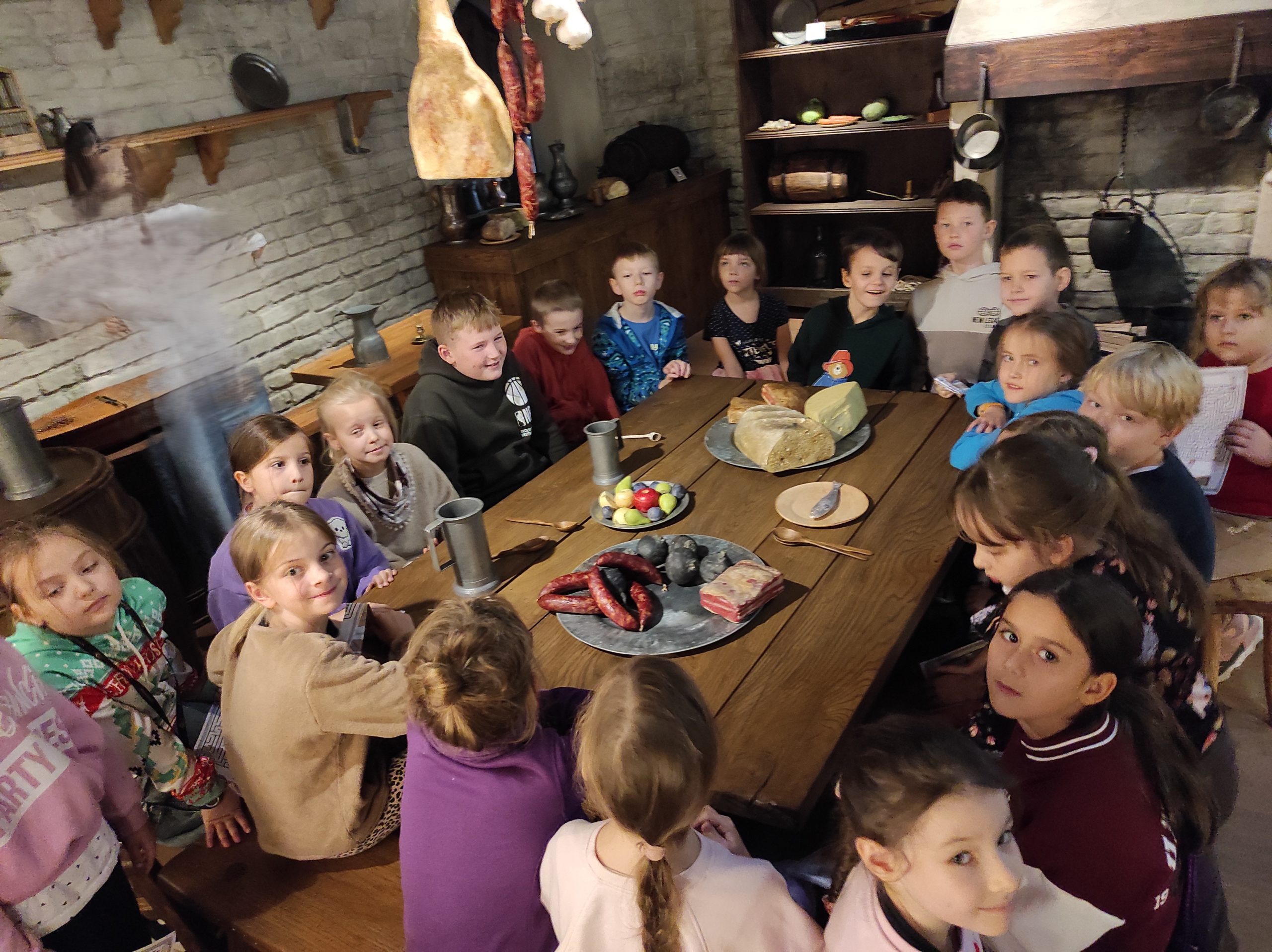 Zdjęcie wykonane w pomieszczeniu. Uczniowie siedzą wokół dużego stołu, na którym leżą rekwizyty przypominające talerze oraz artykuły spożywcze: kiełbasa, boczek, owoce, pieczywo. W tle komoda, regał, ściana.