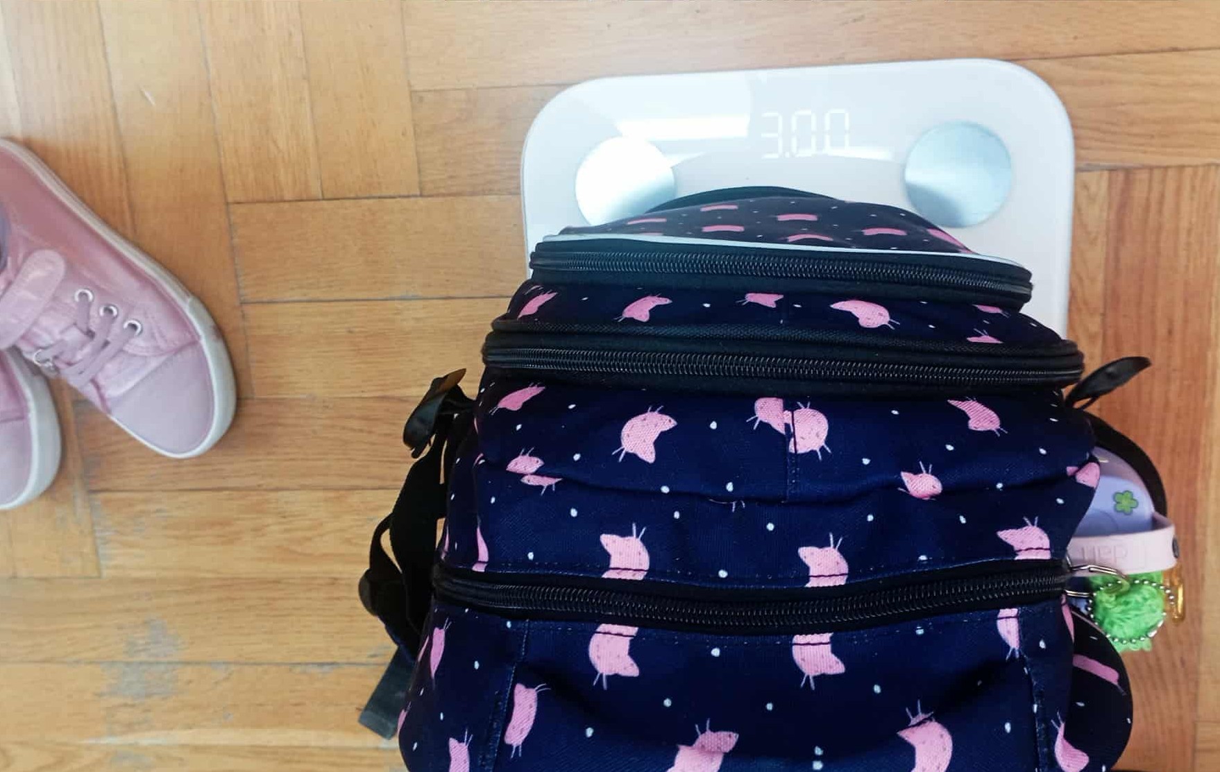 Na wadze elektronicznej, na której wyświetla się 3.00, stoi plecak szkolny. Obok wagi buty dziecięce.