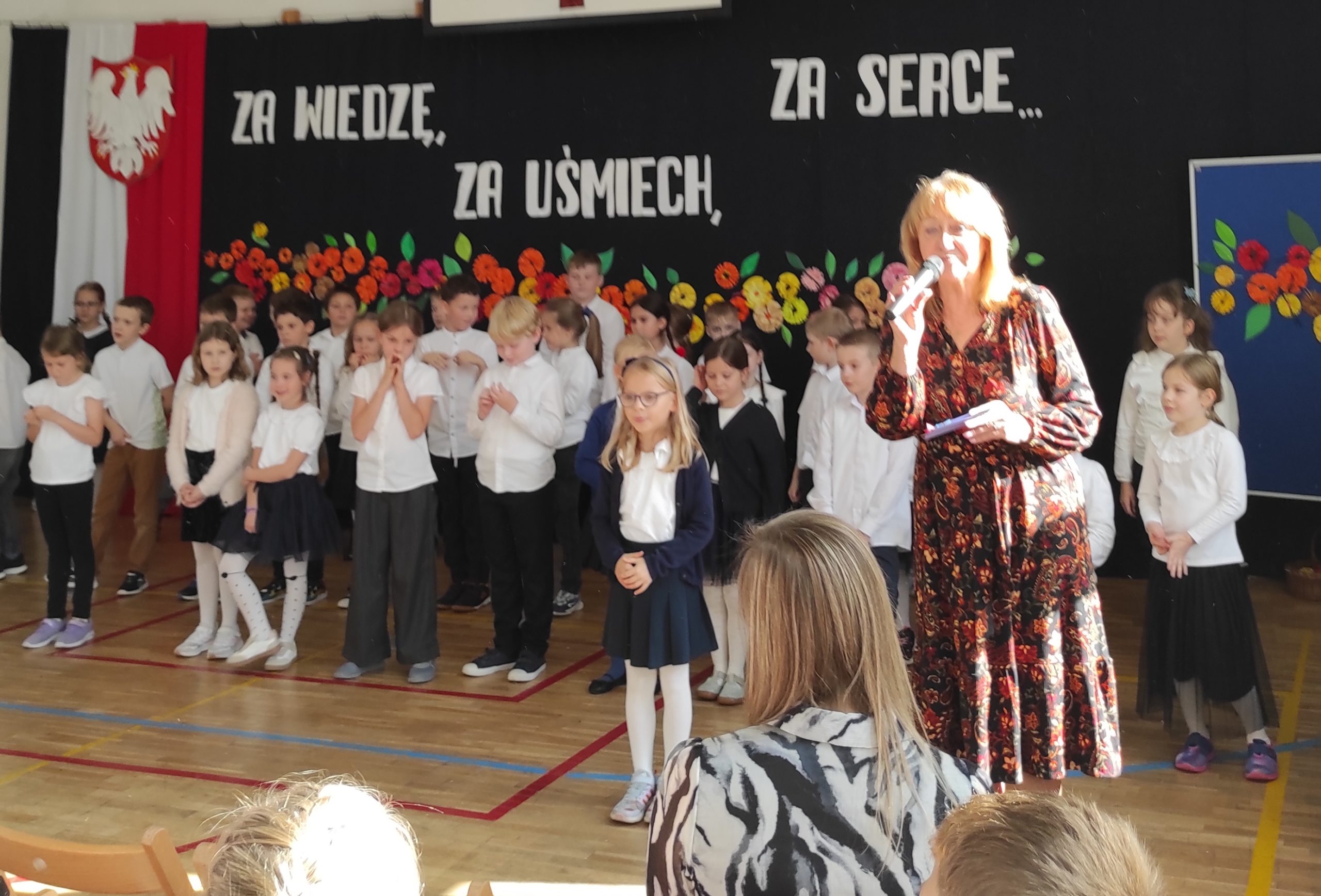 Na pierwszym planie kobieta z mikrofonem w prawej dłoni. Na drugim planie stoją ustawione w trzech rzędach, ubrane na galowo dzieci . W tle dekoracja: zawieszona pionowo flaga Polski, na niej godło,  dalej napis i kwiaty.