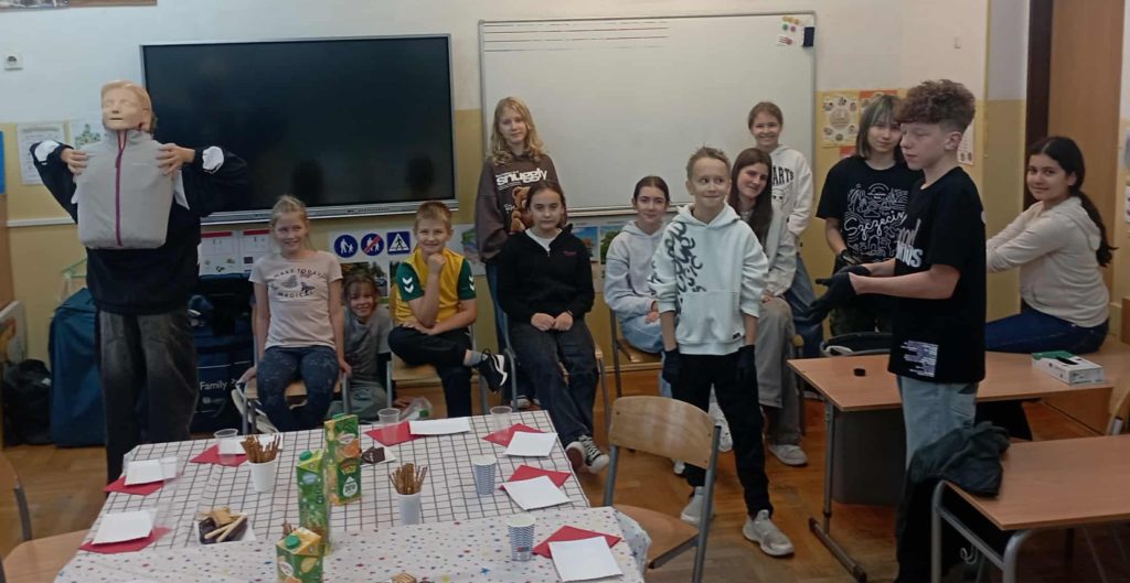 Na te tablic stoją lub siedzą uczniowie Koła PP HOPR, jeden z nich trzyma fantoma. Przed nimi nakryty poczęstunkiem stół.