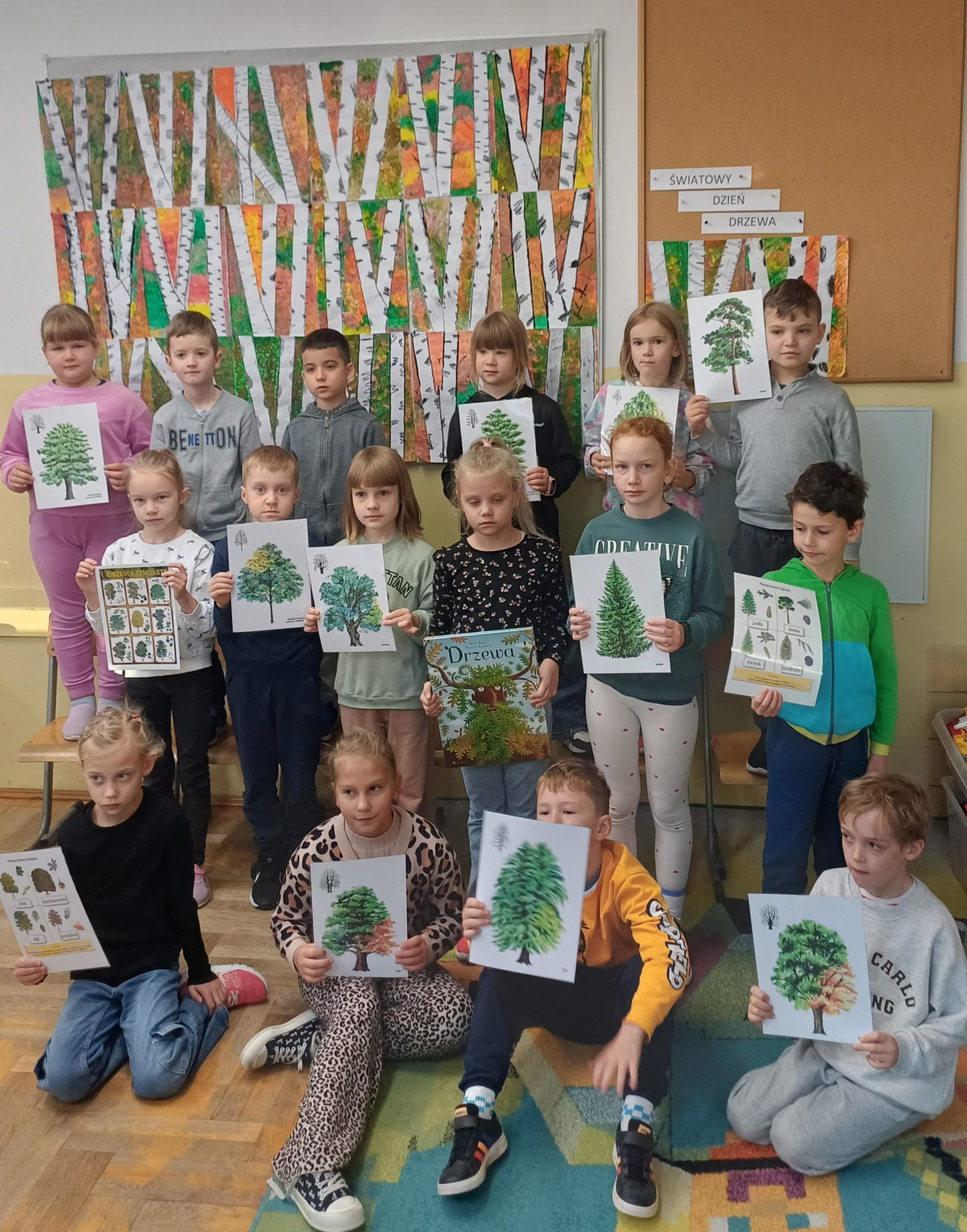 Grupa dzieci trzyma ilustracje drzewa, w tle tablica z rysunkami brzozy.