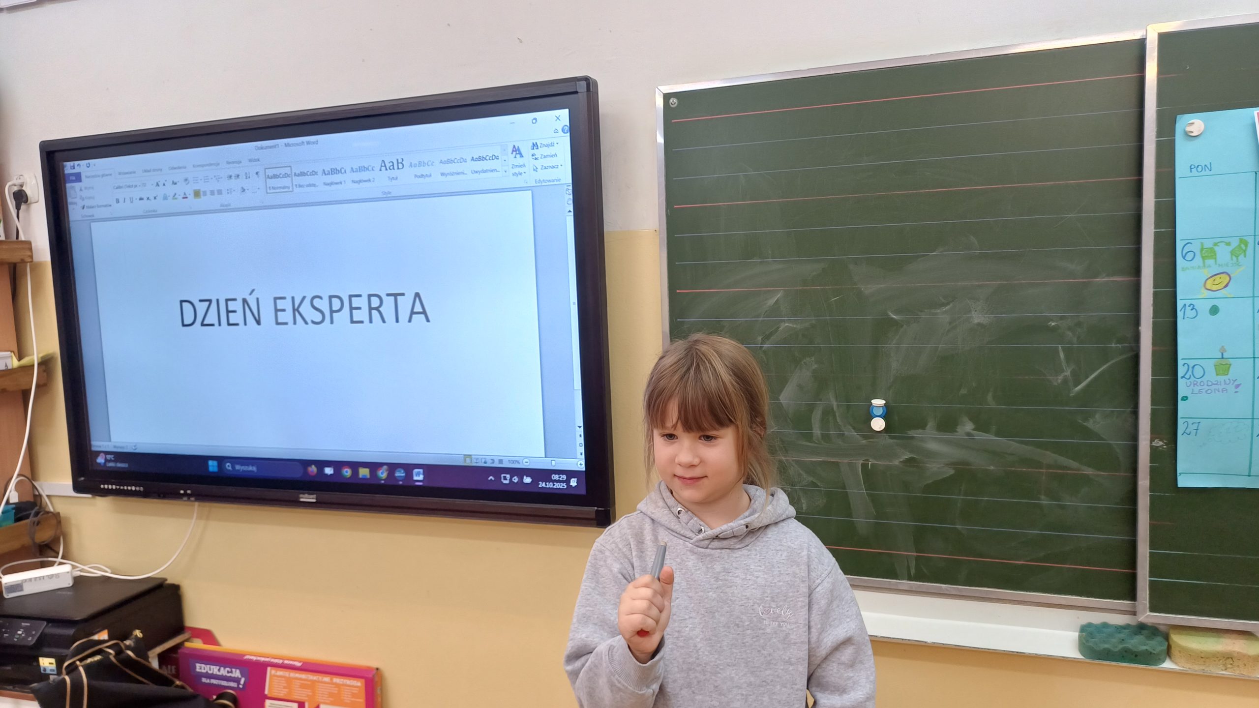 Dziewczynka stoi przed tablicami, w ręku trzymając wskaźnik. Za nią na tablicy napis: "Dzień eksperta".