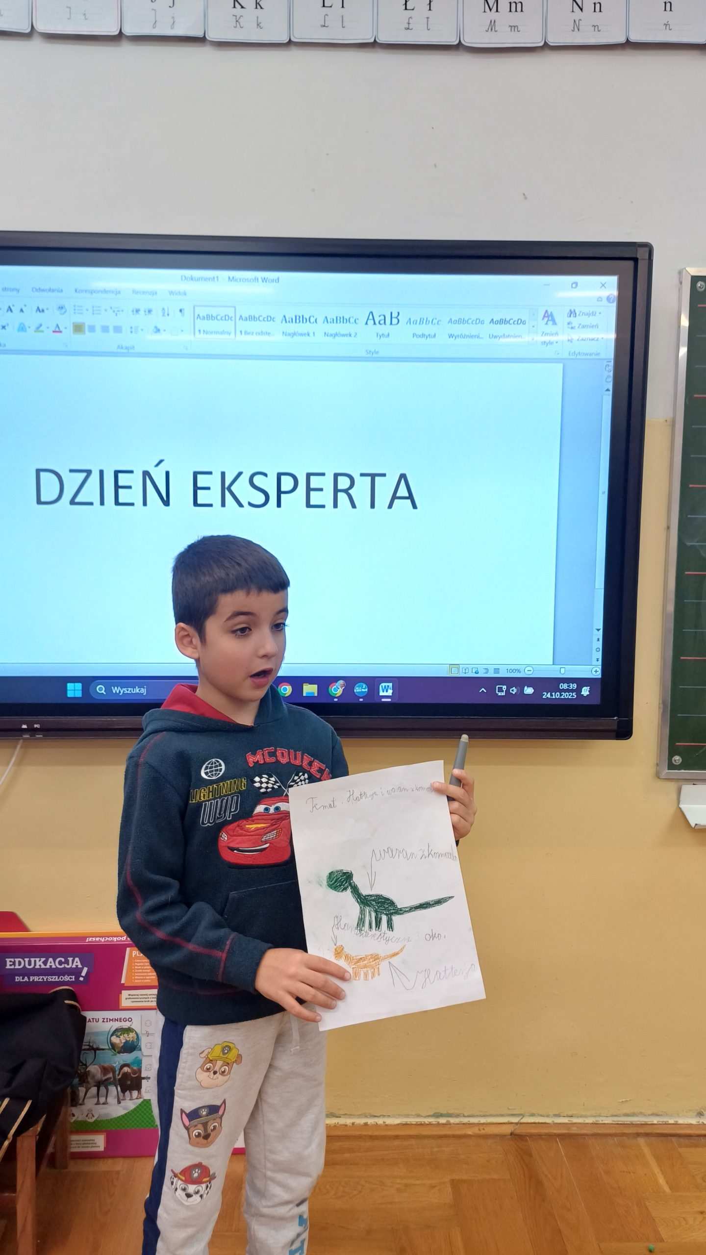 Chłopiec stoi przed tablicą, trzymając w rękach rysunek z dinozaurem