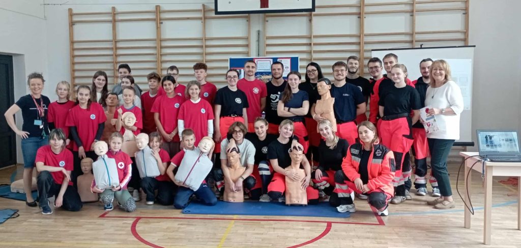 W sali gimnastycznej na tle drabinek i tablicy z plakatem stoją 2 rzędy ludzi oraz 1 rząd przed nimi kuca. Ponad trzydziestoosobowa grupa ludzi pozuje do zdjęcia, część z nich z fantomami w rękach.