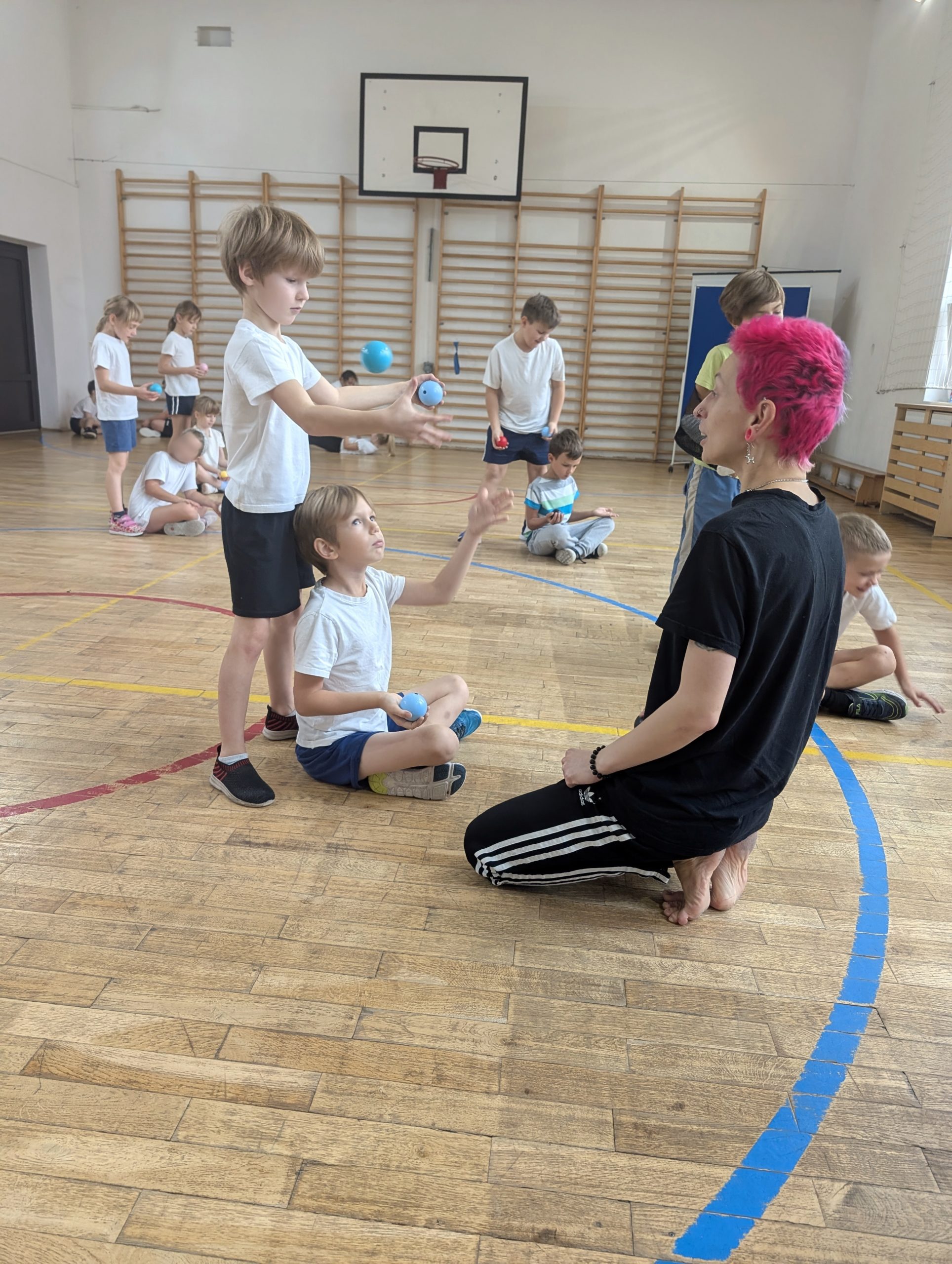 Na pierwszym planie po prawej kuca dorosła osoba i patrzy na 2 chłopców. Jeden siedzi i podrzuca piłeczkę do chłopca stojącego za nim. W tle inni uczniowie wykonujący to samo ćwiczenie.