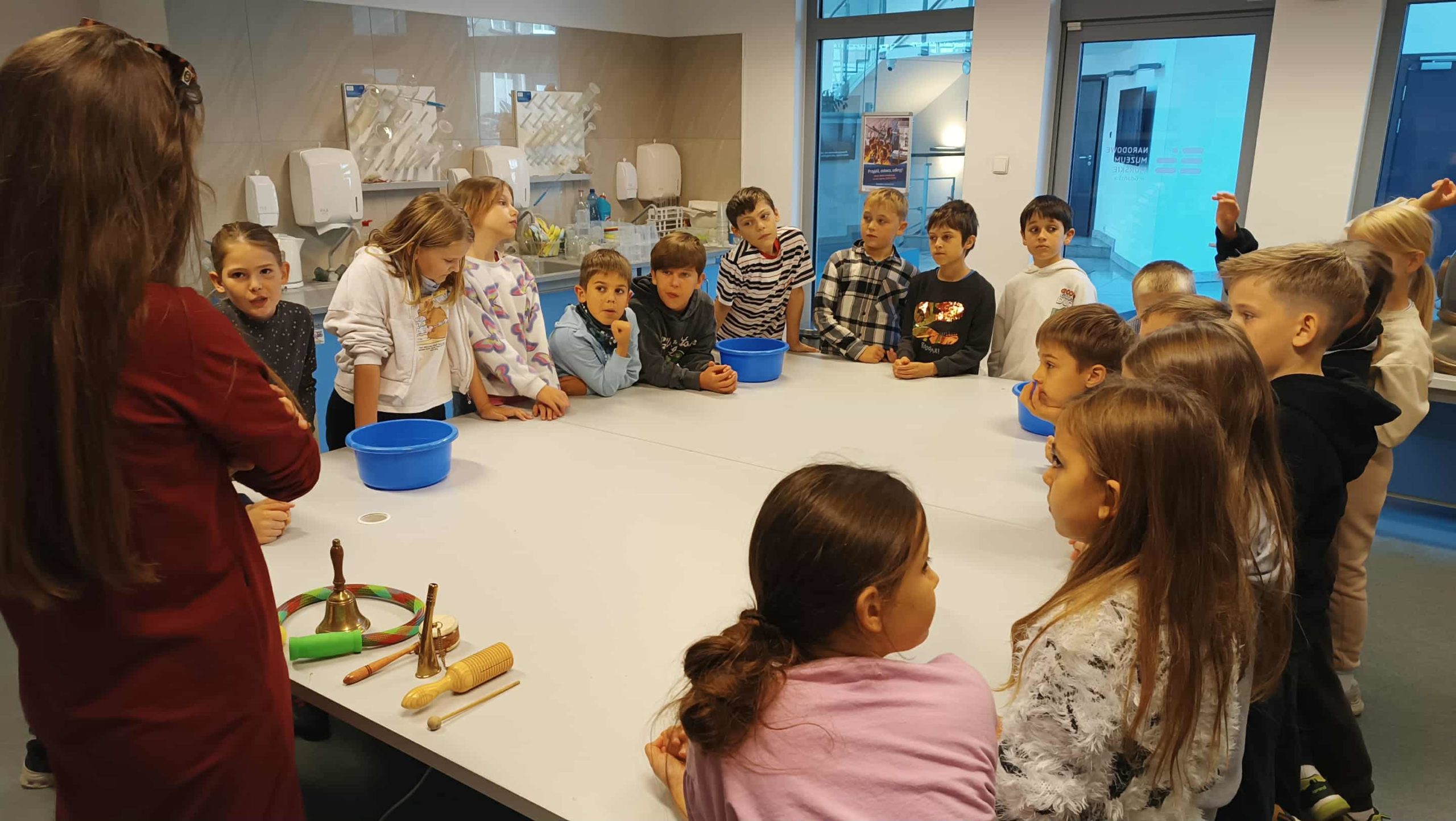 Pani prowadząca warsztaty rozmawia z grupą uczniów klasy 3c stojących wokół  dużego kwadratowego stołu  w muzeum.