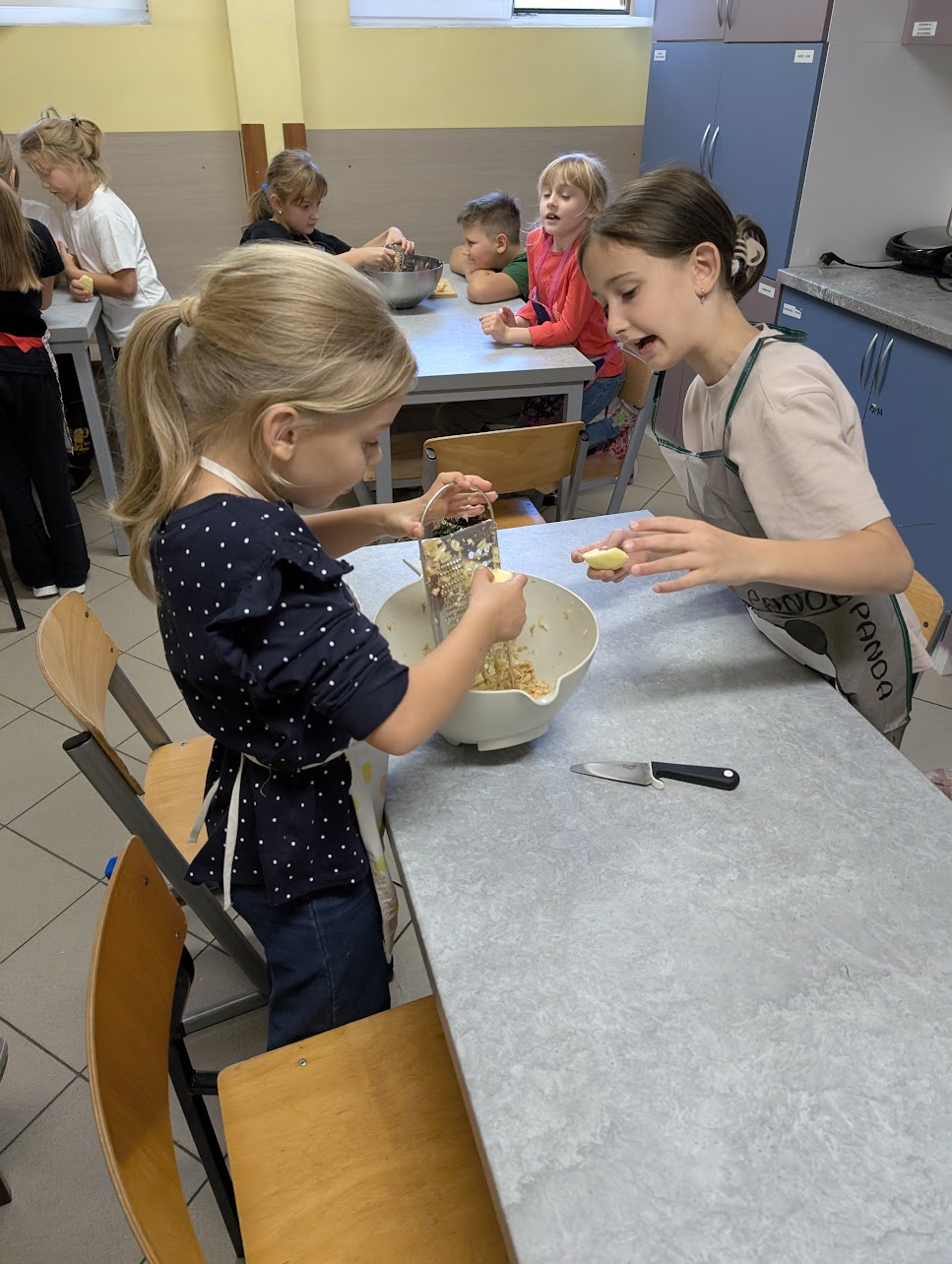 Grupa dzieci i kobieta przygotowująca wypieki w kuchni lub sali warsztatowej. Na stole leżą surowe ciasteczka lub bułeczki ułożone na blasze wyłożonej papierem do pieczenia. Kobieta w okularach i niebieskim fartuchu rozsmarowuje farsz. Dzieci w różnym wieku uśmiechają się i pozują do zdjęcia – niektóre pokazują znak pokoju, inne patrzą z zaciekawieniem. W tle widać ławkę i ścianę w jasnych kolorach.