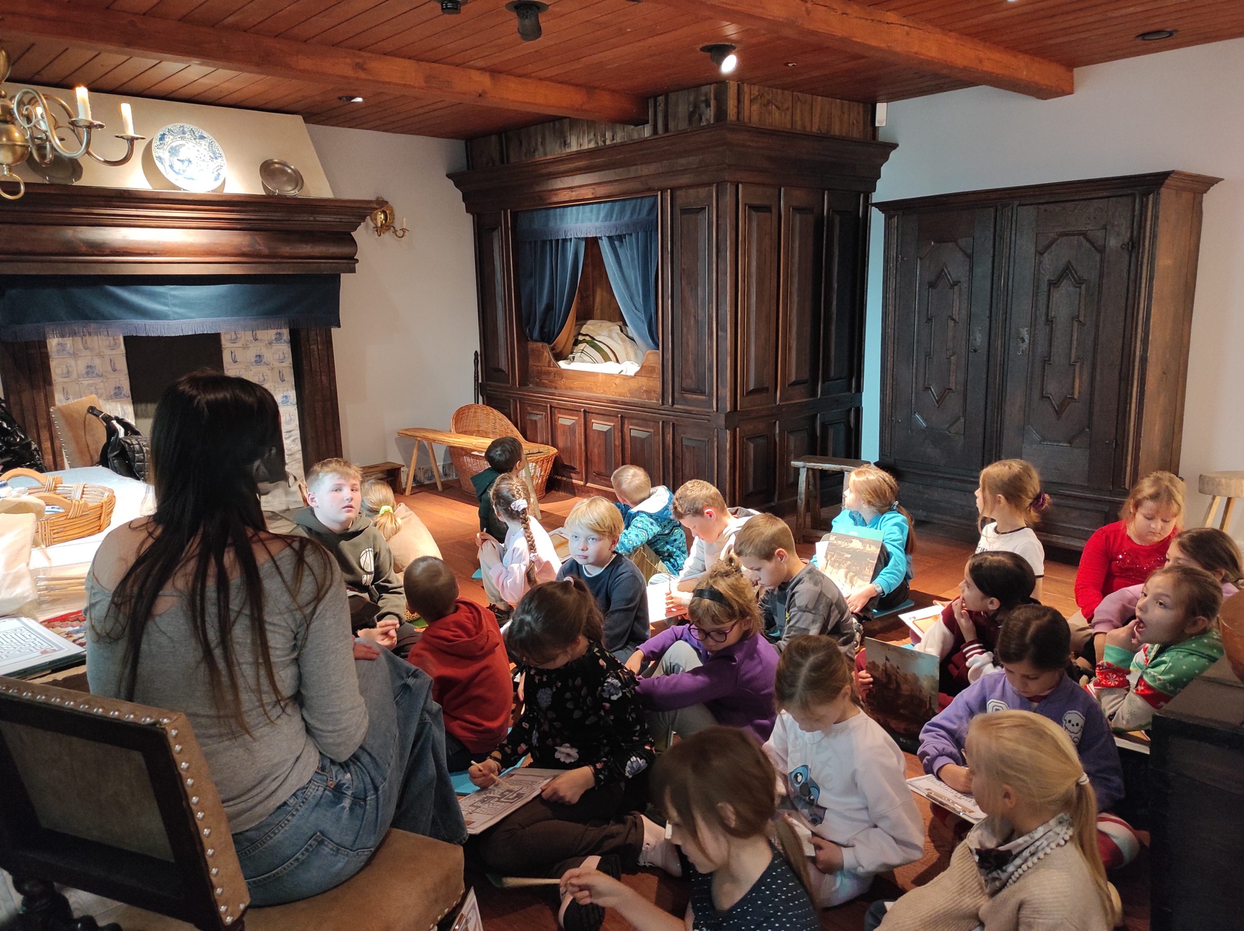 Zdjęcie wykonane w pomieszczeniu. Dzieci siedzą na podłodze. W tle wyposażenie pokoju: ozdobna, drewniana szafa, zabudowane łózko, wiklinowe łóżeczko dla dziecka, fragment pieca kaflowego i drewnianego sufitu.