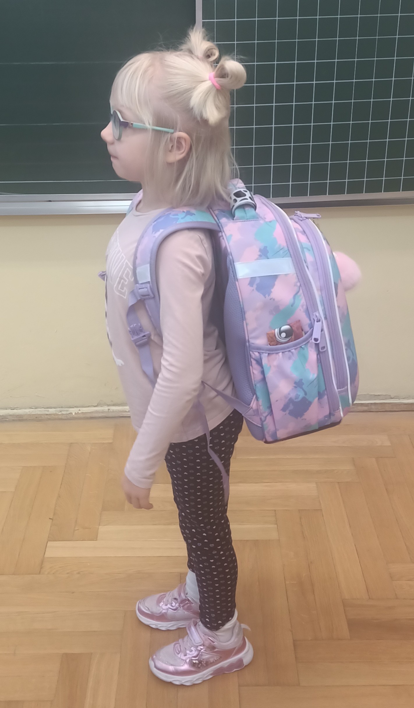 Na tle tablicy szkolnej stoi wyprostowana dziewczynka z plecakiem na plecach.