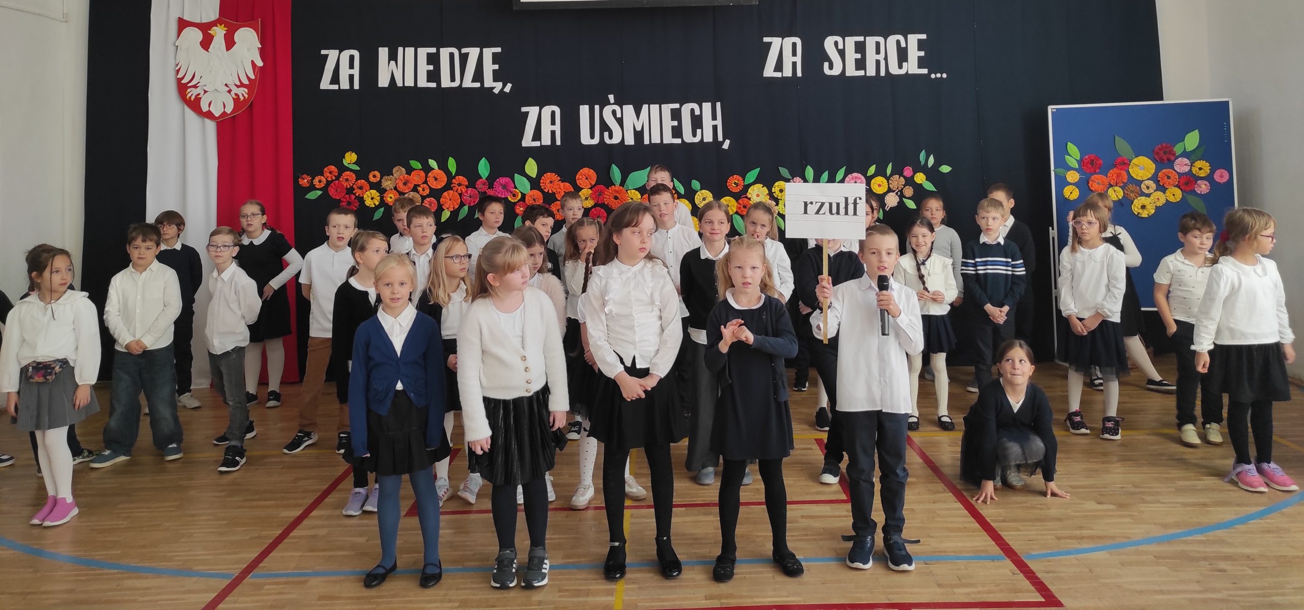 Na pierwszym planie stoi 5 uczniów, jeden z nich trzyma  w prawej dłoni napis "rzułf", a w lewej mikrofon. Za nimi stoją  ustawione w trzech rzędach, ubrane na galowo dzieci.  W tle dekoracja: zawieszona pionowo flaga Polski, na niej godło,  dalej napis i kwiaty.