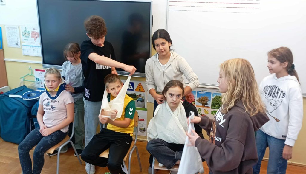 Na tle tablic siedzi troje uczniów. W koło nich inni członkowie Koła PP HOPR zakładający temblaki na ręce lub kołnierz na szyję.