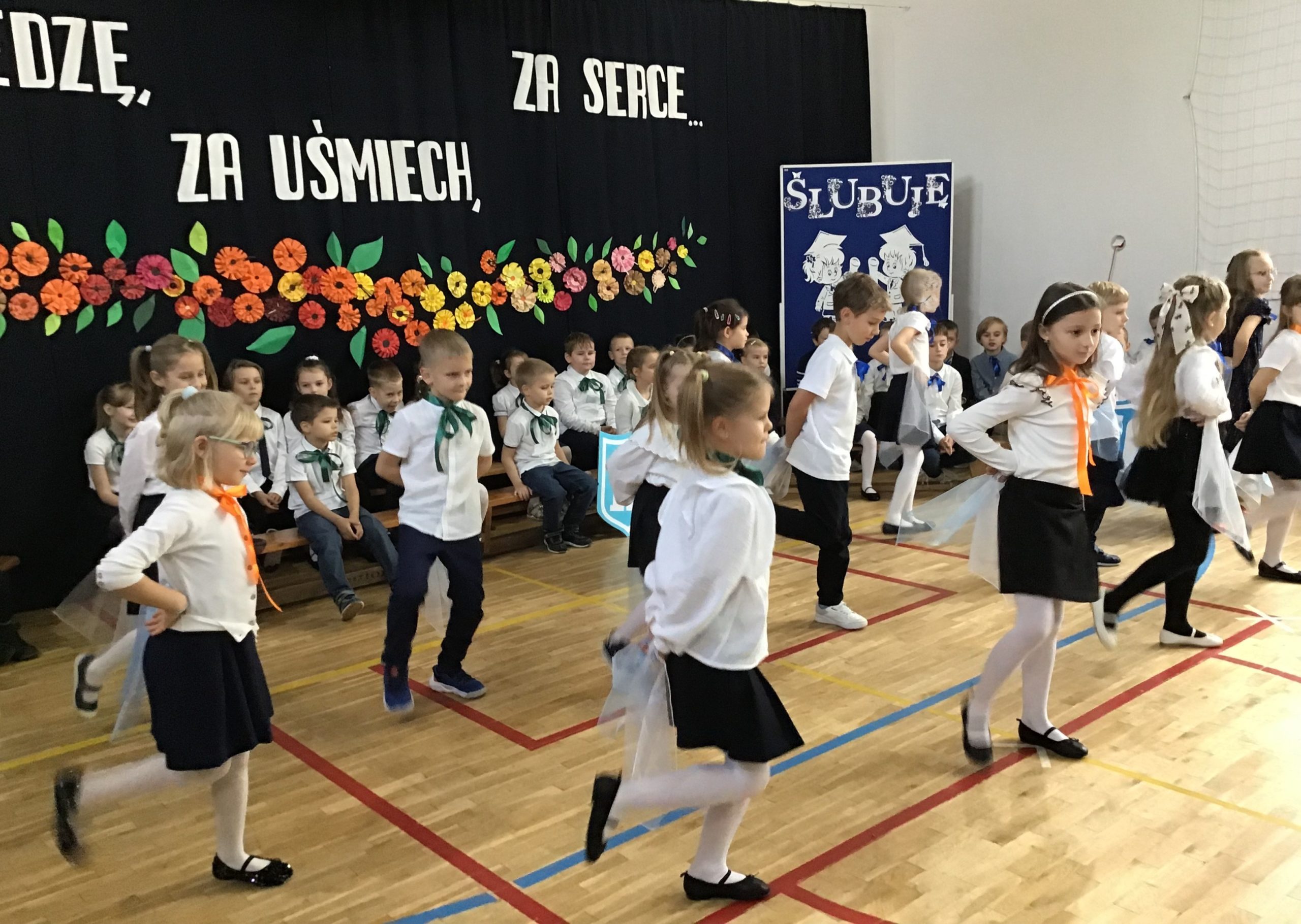 Na środku sali gimnastycznej w trzech rzędach stoi po 5 reprezentantów klas pierwszych wymachując chustkami trzymanymi w rękach. W tle siedzą pozostali uczniowie klas pierwszych.