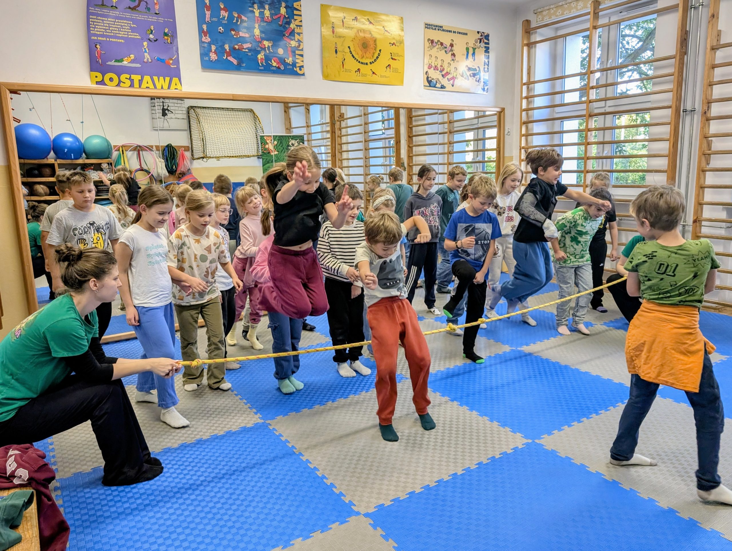 Grupa dzieci uczestniczy w zabawie ruchowej w sali gimnastycznej. Dzieci przeskakują przez rozciągniętą na wysokości liny, którą trzymają dwie osoby – jedna z lewej strony w zielonej koszulce i druga z prawej w zielonej koszulce z pomarańczową bluzą przewiązaną w pasie. Pozostałe dzieci stoją w kolejce, czekając na swoją kolej.