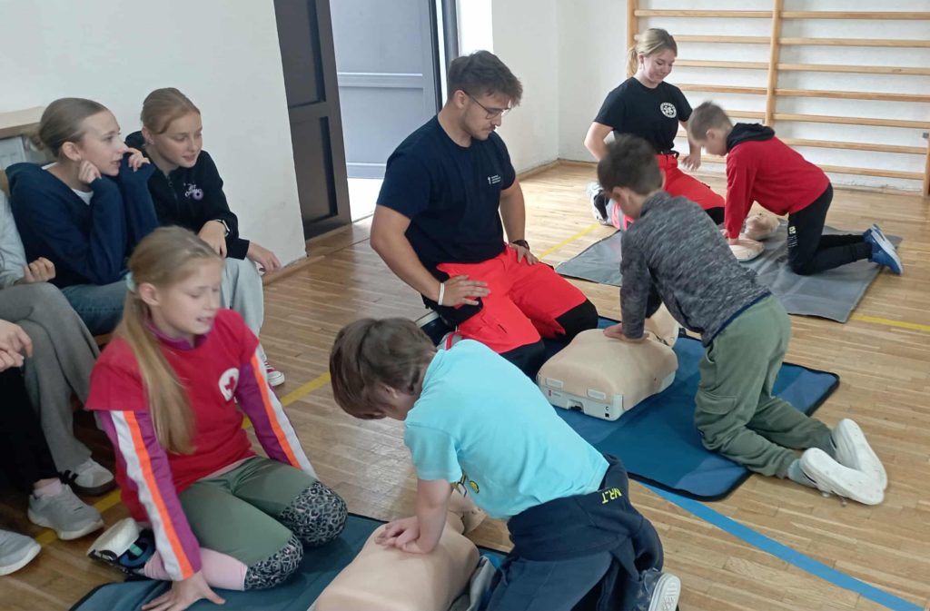Trzech młodszych uczniów uciska klatkę piersiową leżących fantomów. W tle 2 ratowników i uczennica koła PP oraz dwie dziewczynki.