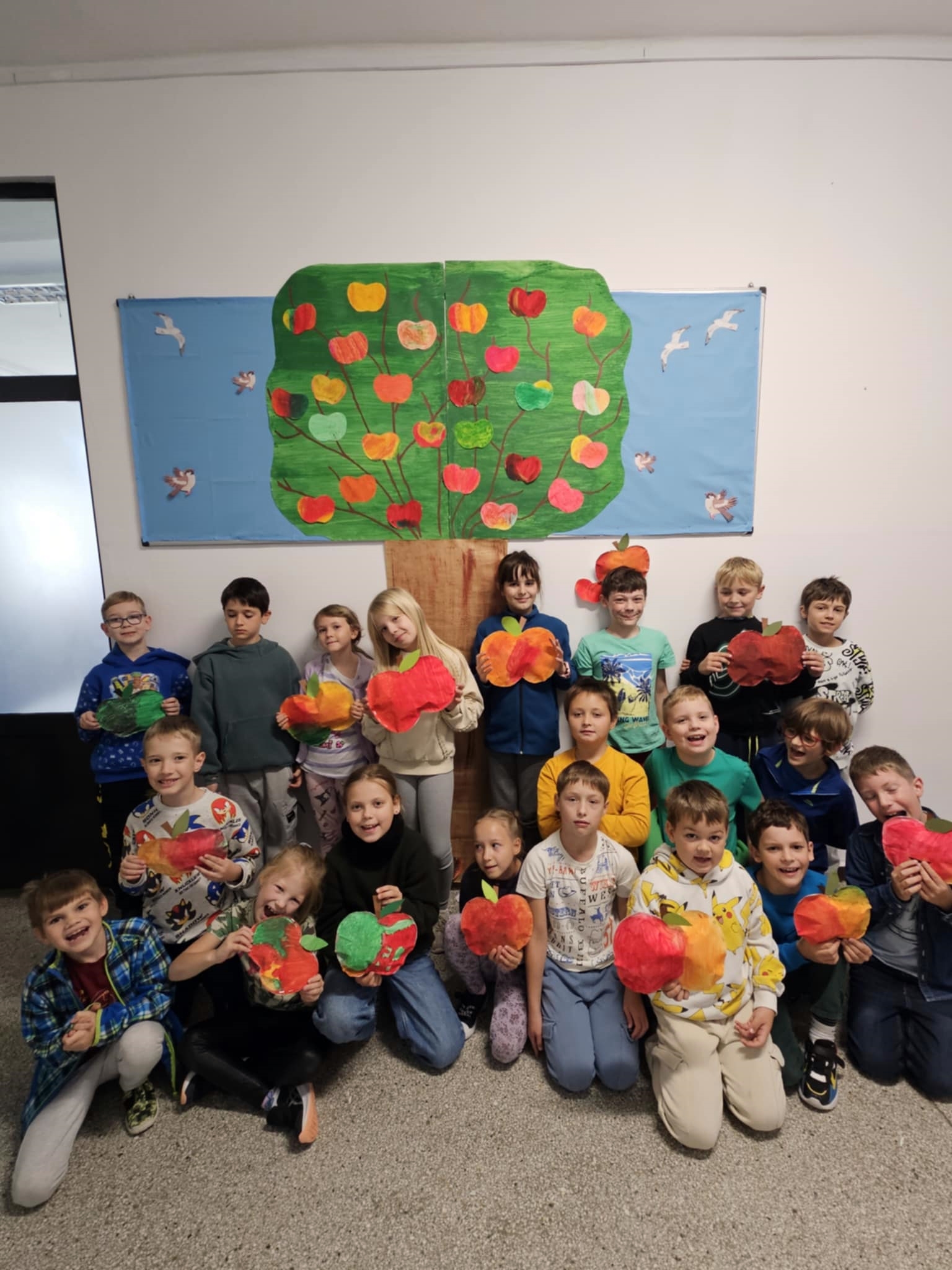 Duża grupa uczniów stoi pod papierowym drzewem z kolorowymi jabłkami, trzymając w rękach własnoręcznie wykonane prace plastyczne w kształcie jabłek.