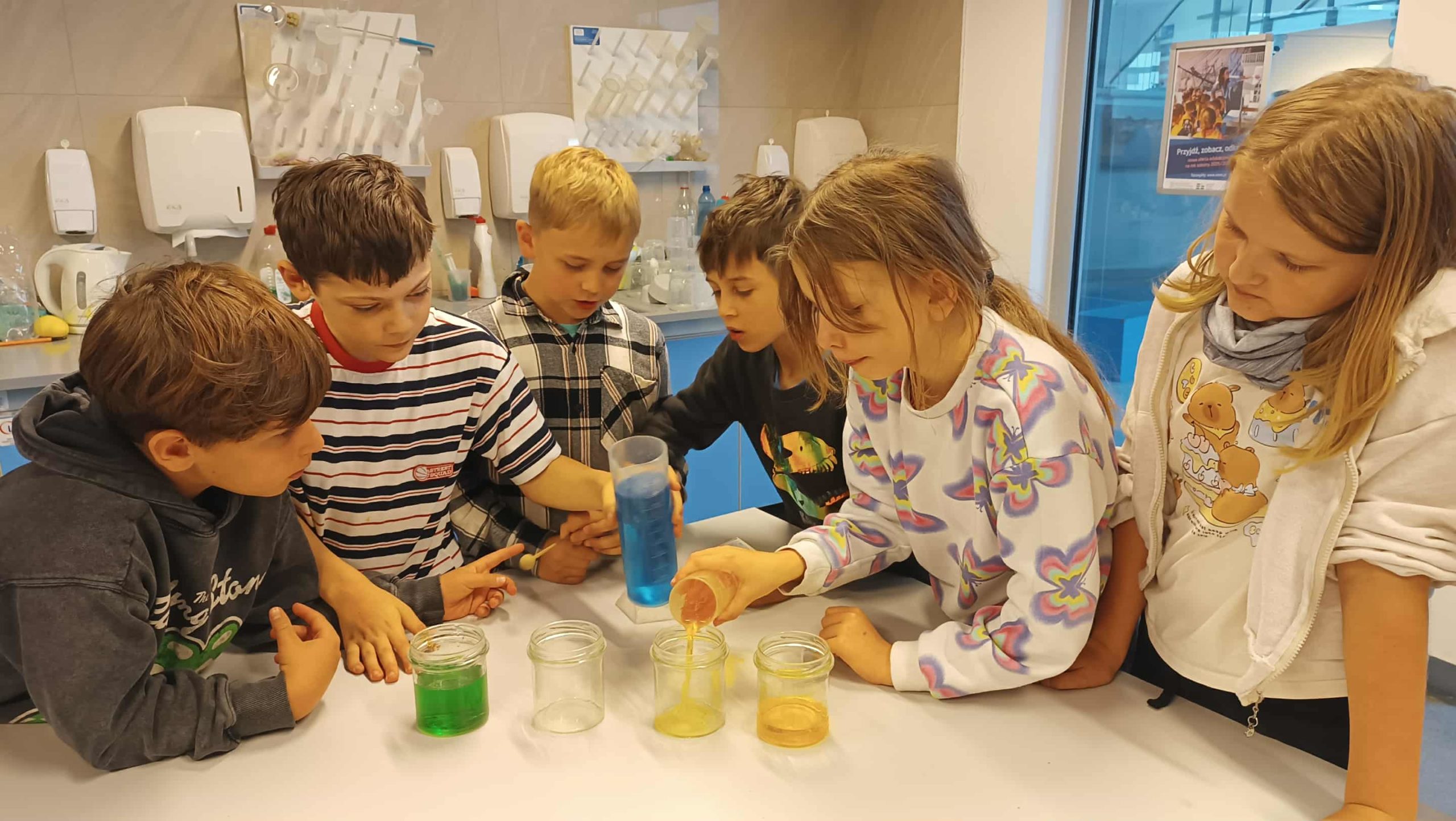 Dzieci z klasy 3c przelewają kolorowe płyny do słoików, wykonując doświadczenia podczas zajęć „Muzyka na wodzie”.