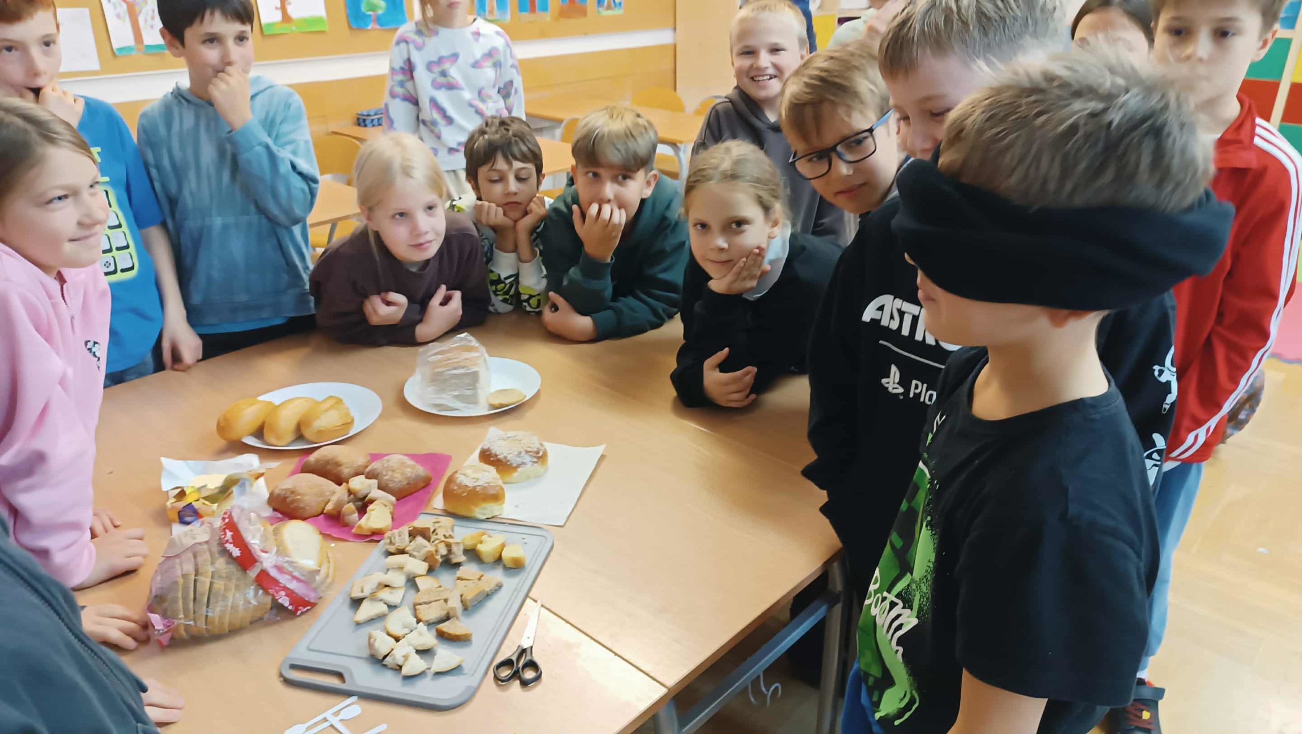  Uczniowie w klasie obserwują kolegę, który z zasłoniętymi oczami rozpoznaje różne rodzaje pieczywa podczas zajęć o chlebie