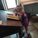 Zdjęcie wykonane w pomieszczeniu. Na pierwszym planie siedząca na krześle, przy stoliku dziewczynka. Na stole leży otwarty notes. Twarz dziecka skierowana jest w jego stronę. W prawej dłoni trzyma ptasie pióro.