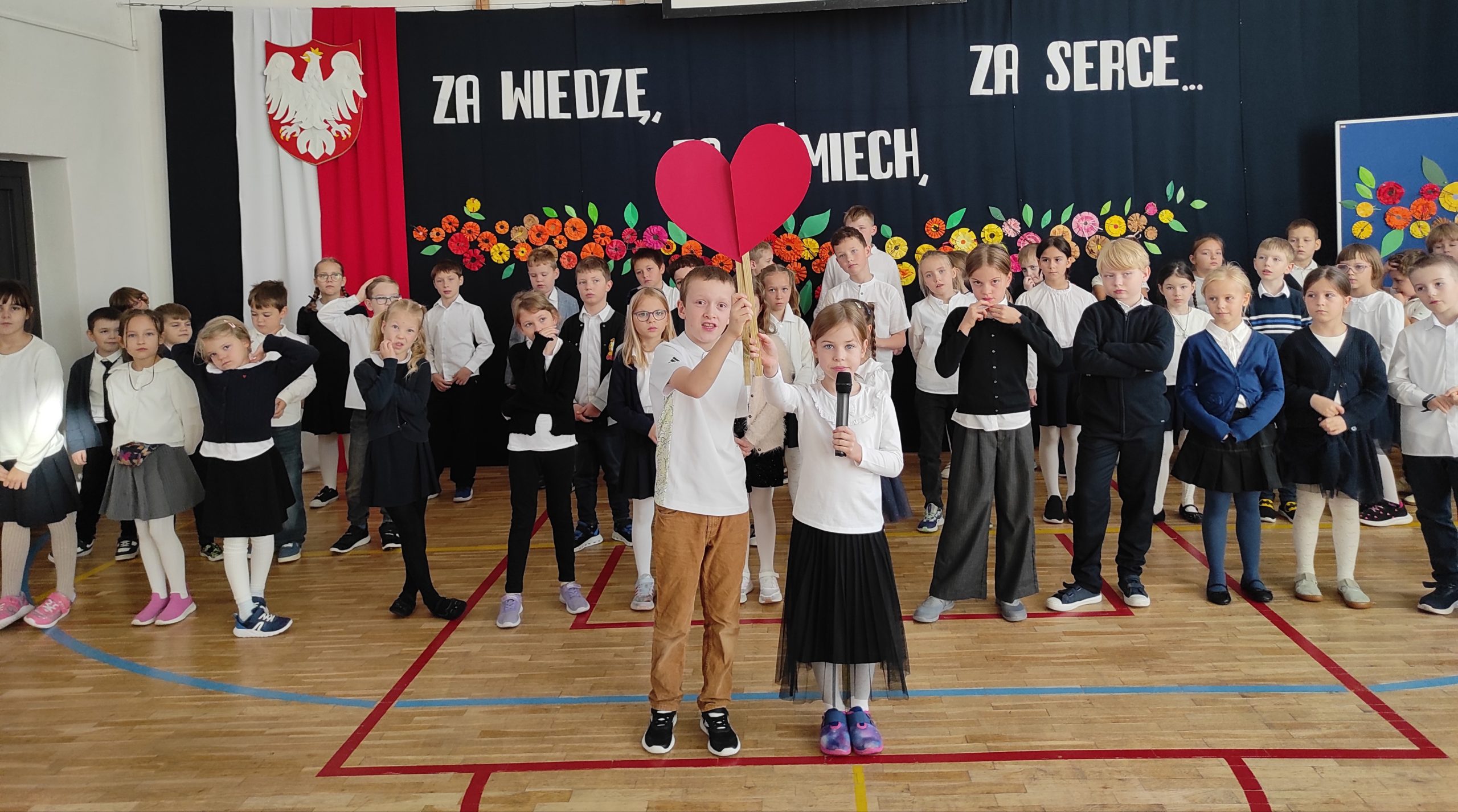 Na pierwszym planie stoi dwoje uczniów, dziewczynka i chłopiec. Każde z nich trzyma w dłoni patyk, do którego doczepiona jest połowa serca.  Łączą je w całość. Za nimi stoją  ustawione w trzech rzędach, ubrane na galowo dzieci. W tle dekoracja: zawieszona pionowo flaga Polski, na niej godło,  dalej napis i kwiaty.