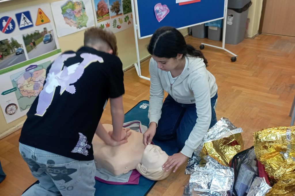 Przy fantomie uczeń uciskający klatkę piersiową oraz uczennica udrażniająca drogi oddechowe. Obok nich rozwinięte koce ratownicze.