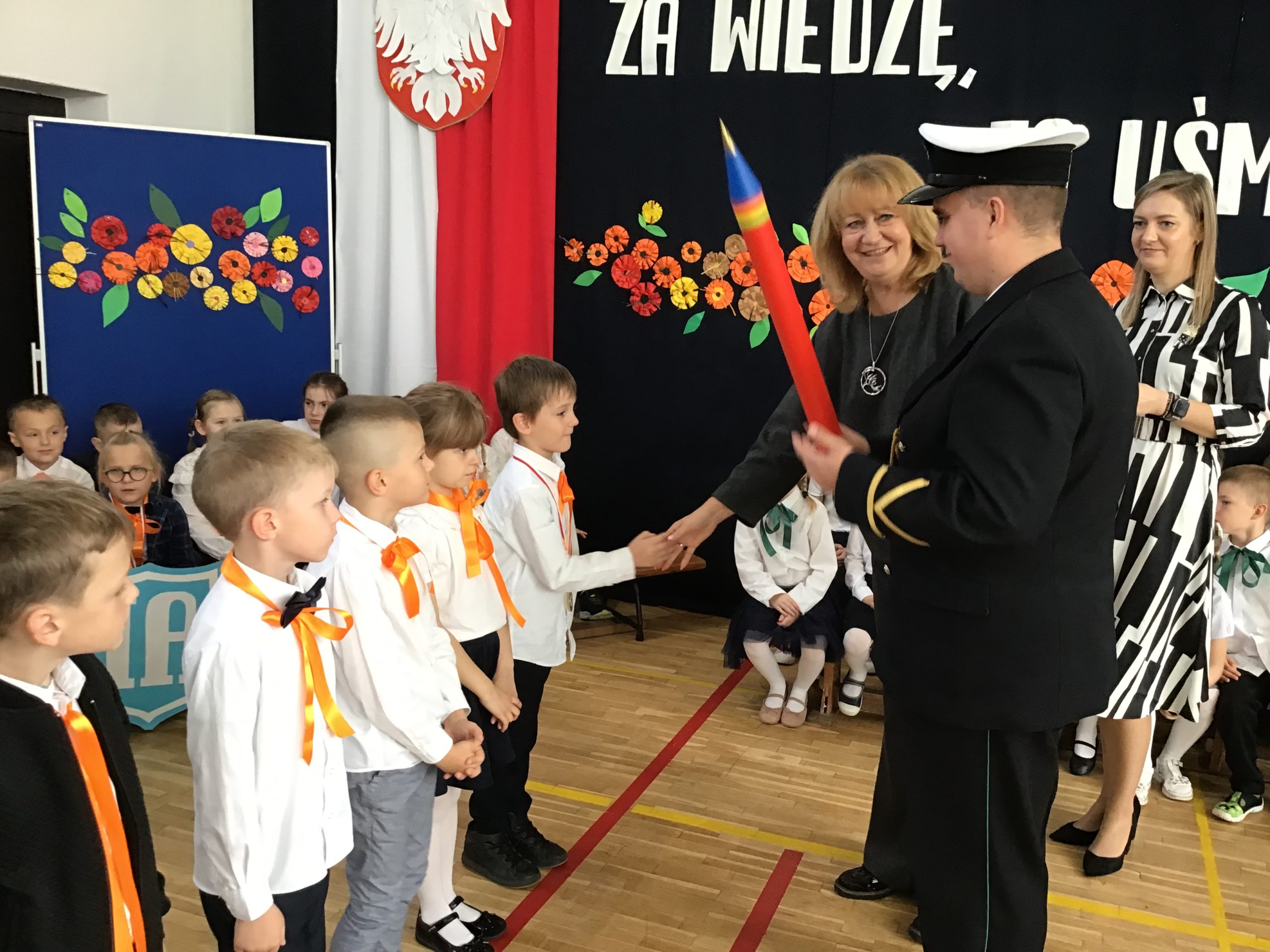 Na środku sali gimnastycznej Pani Dyrektor ściska rękę pasowanego ucznia. Na pierwszym planie funkcjonariusz Straży Granicznej w galowym stroju. Obok ucznia stoi czwórka innych uczniów klasy IA. W tle pozostali uczniowie i dekoracja kwiatowa.