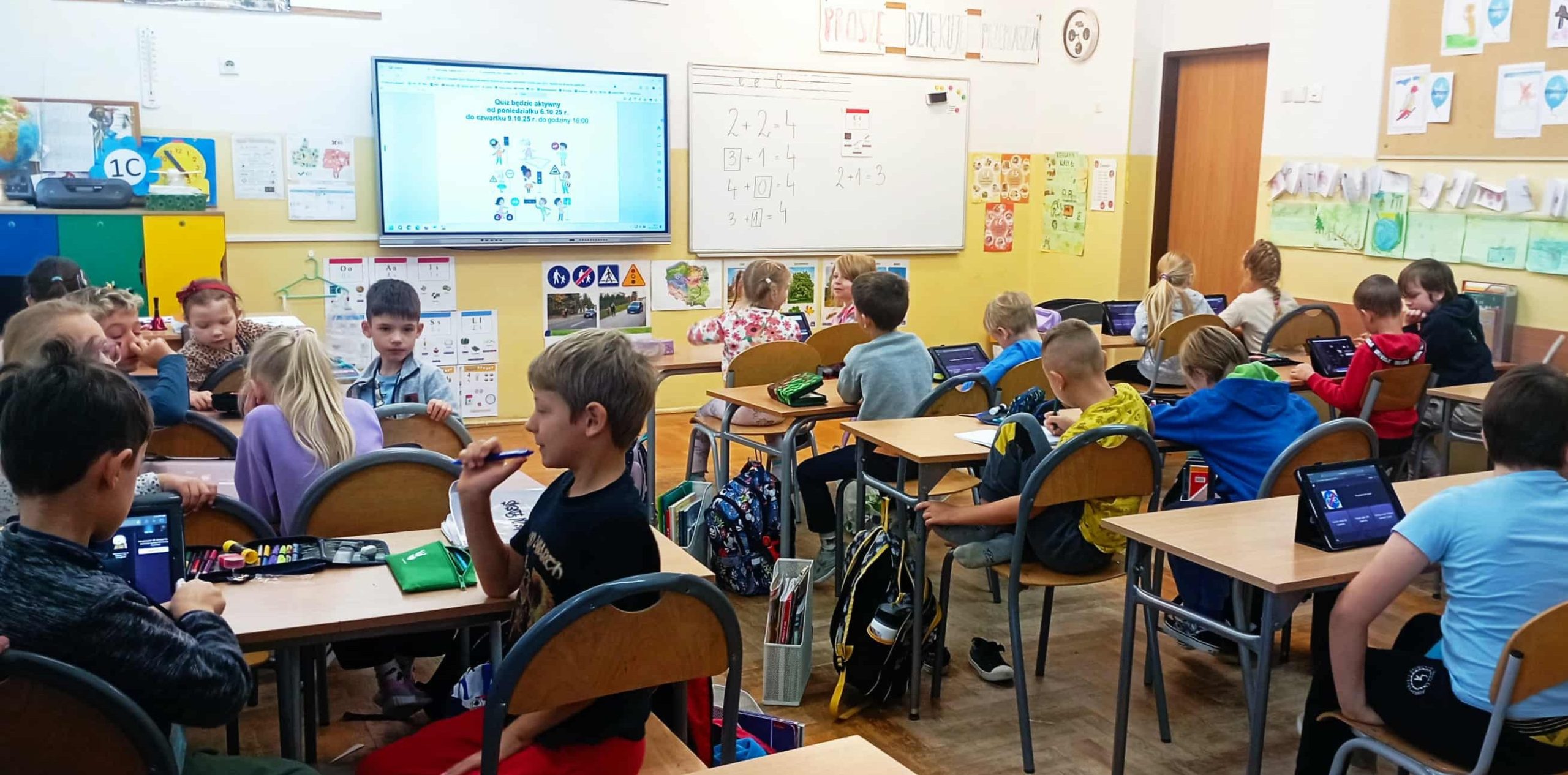 W sali lekcyjnej w trzech rzędach ławek siedzą obróceni tyłem uczniowie klasy 1c. Przed uczniami stoją na ławce IPady, na którym wyświetlają się pytania quizu. W tle tablica multimedialna z plakatem konkursu „Bezpieczna droga do szkoły”