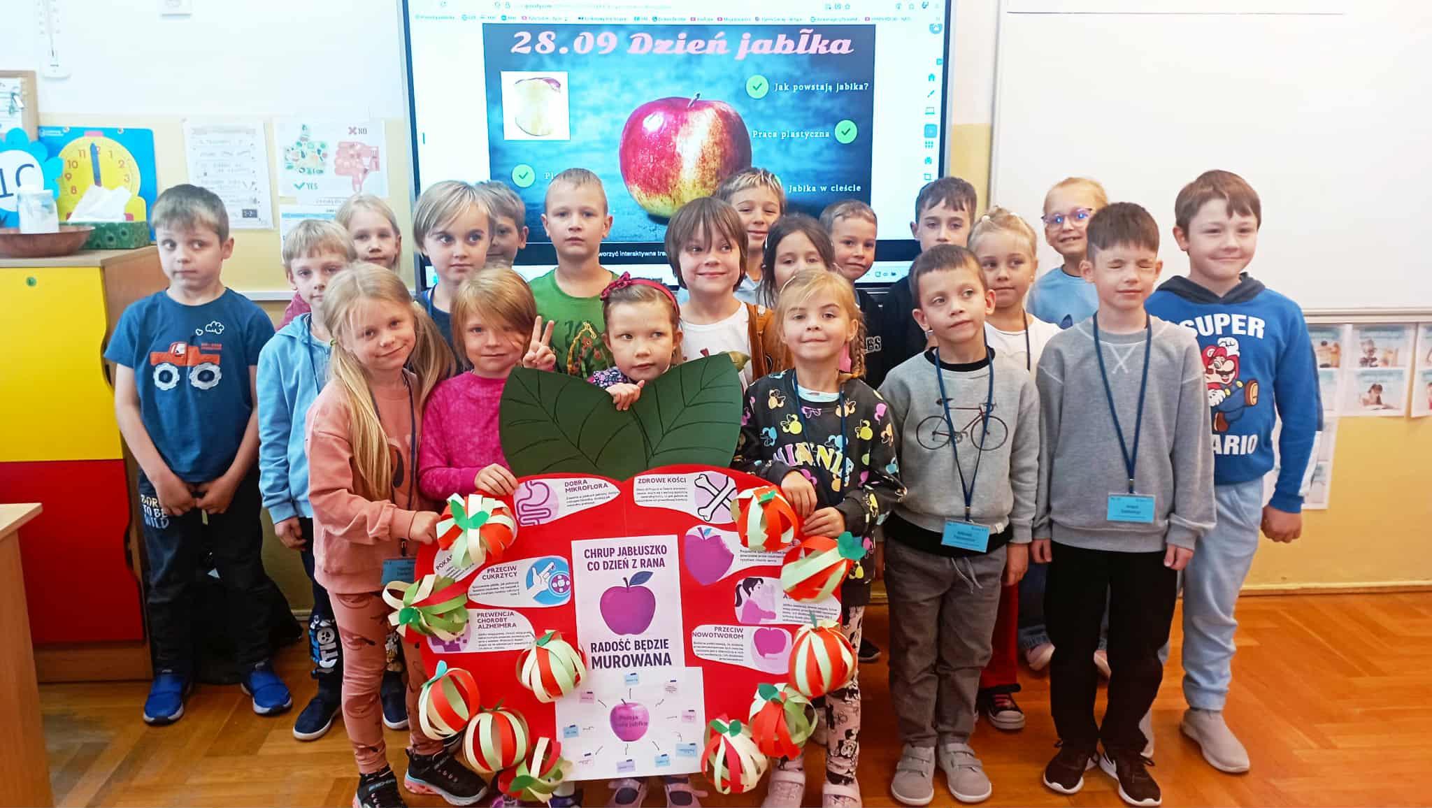 Grupa uczniów pozuje w klasie z dużym, kolorowym plakatem w kształcie jabłka, zawierającym hasła promujące zdrowe odżywianie. W tle tablica z napisem „28.09 Dzień Jabłka".