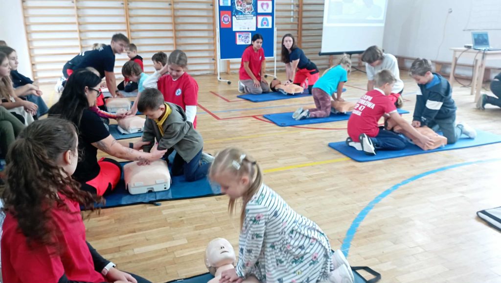 W sali gimnastycznej przy 8 fantomach klęczą po 2 osoby. Jedna z nich uciska klatkę fantoma. W tle tablica z plakatem RKO i ekran rzutnika.