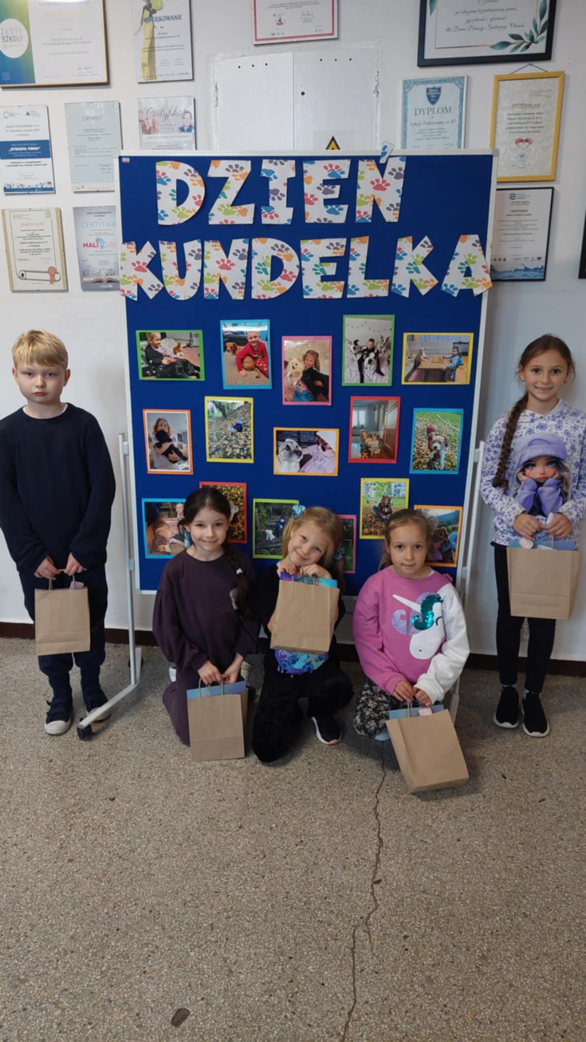 Grupa pięciorga dzieci stoi przed kolorową tablicą z napisem „Dzień Kundelka”, ozdobioną zdjęciami uczniów z psami. Dzieci trzymają w dłoniach papierowe torebki z nagrodami i uśmiechają się do aparatu.