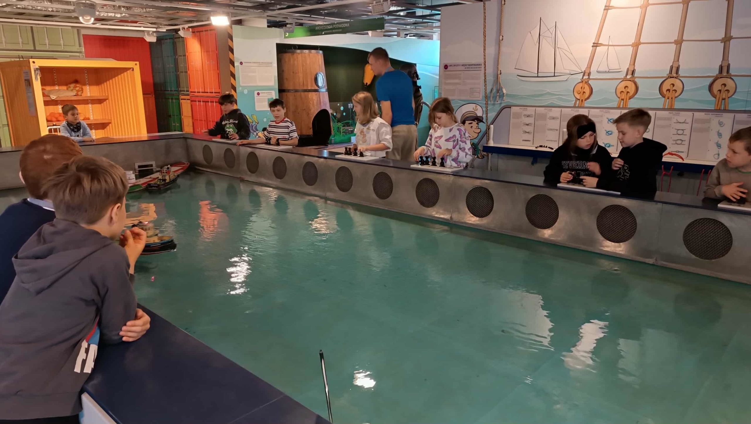 Dzieci sterują miniaturowymi statkamiw dużym basenieedukacyjnym, ucząc się zasad żeglugi i manewrowania jednostkami.