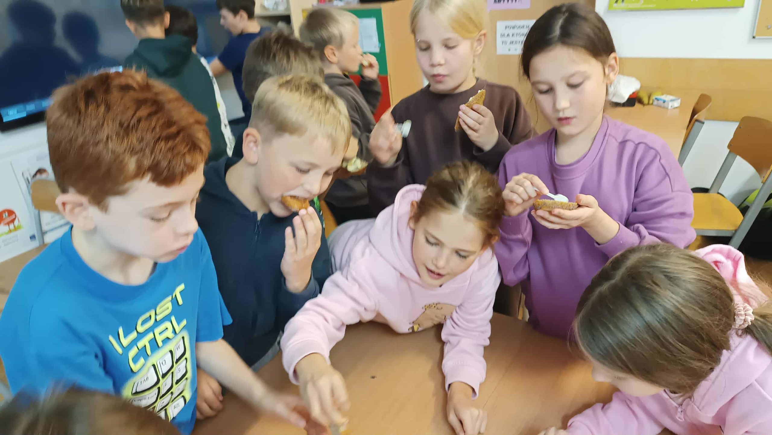  Uczniowie próbują różnych rodzajów pieczywa, rozpoznając smaki z zamkniętymi oczami. Na stole widać pieczywo i talerze, dzieci są skupione i uśmiechnięte.