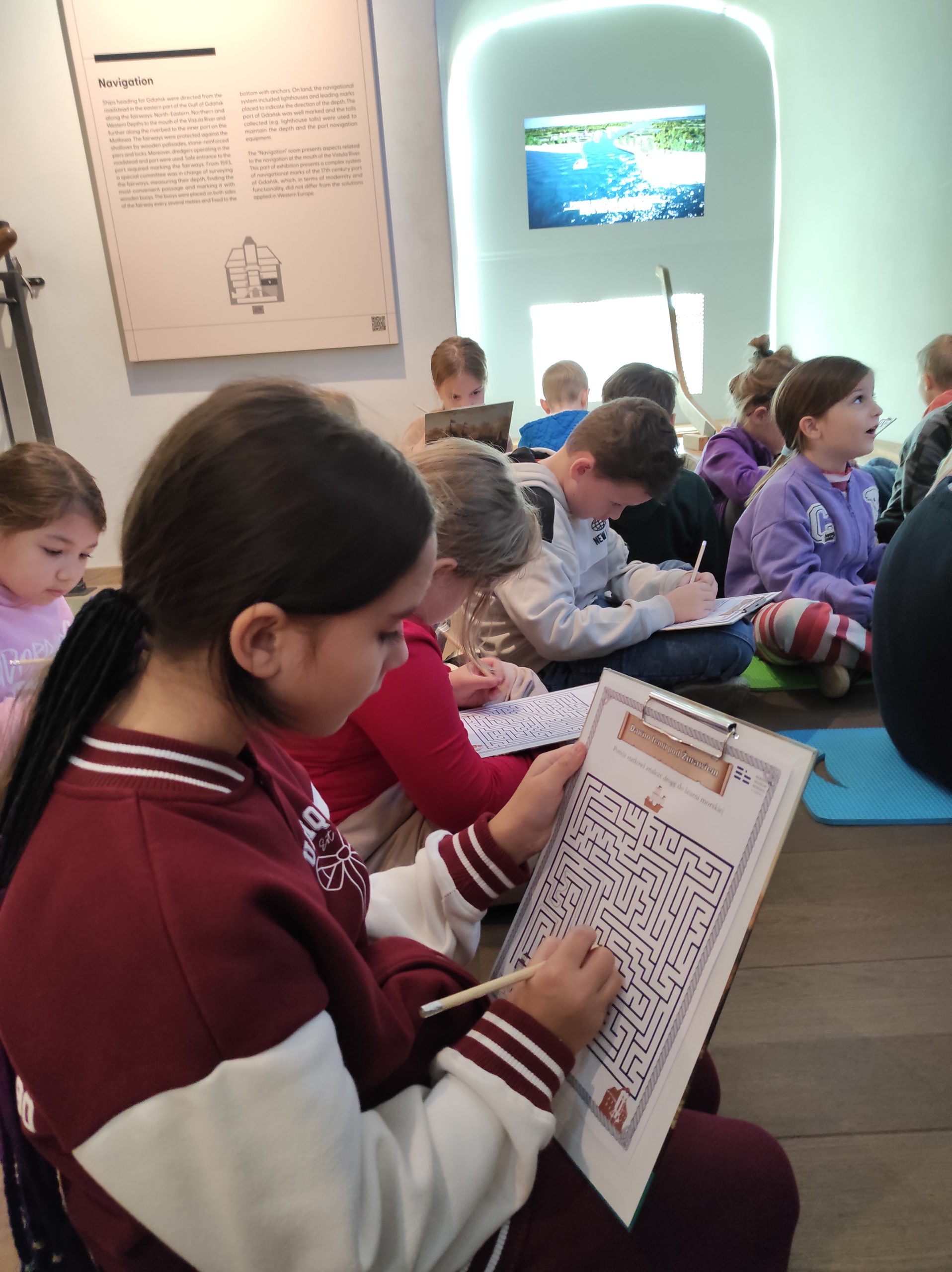 Zdjęcie wykonane w pomieszczeniu. Kilkoro uczniów siedzi na podłodze. Na pierwszym planie dziewczynka, która trzyma lewą dłonią podkładkę z wydrukowaną kartką, a w prawej ołówek. W tle inne dzieci, ściana i wyposażenie sali. 