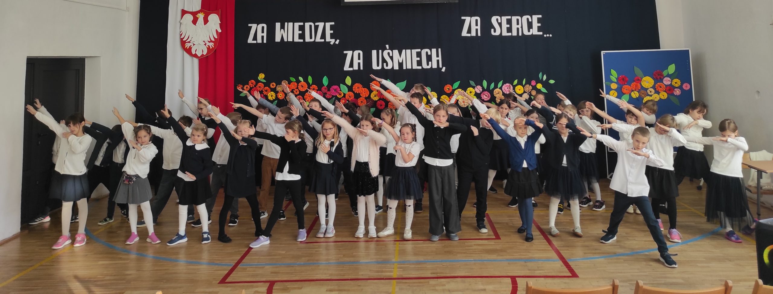 Na pierwszym planie stoją  ustawione w trzech rzędach, ubrane na galowo dzieci. Wszyscy mają podniesione i skierowane w prawą stronę ręce. W tle dekoracja: zawieszona pionowo flaga Polski, na niej godło,  dalej napis i kwiaty.