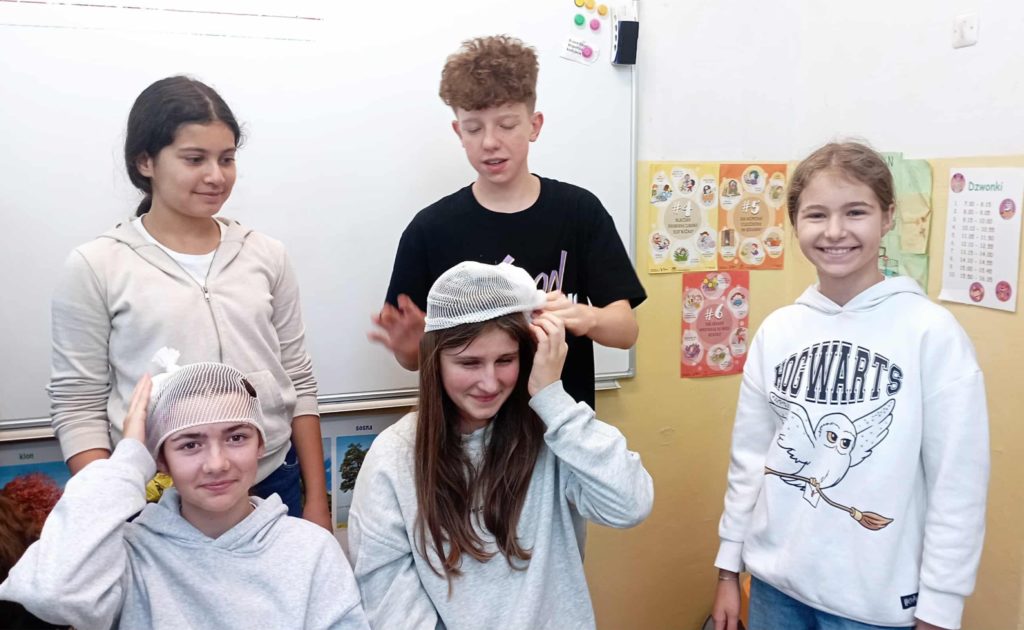 Na pierwszym planie dwie siedzące uczennice, którym pozostała 3 Koła HOPR zakłada opatrunek na głowę z gazy i siatki Codofix.