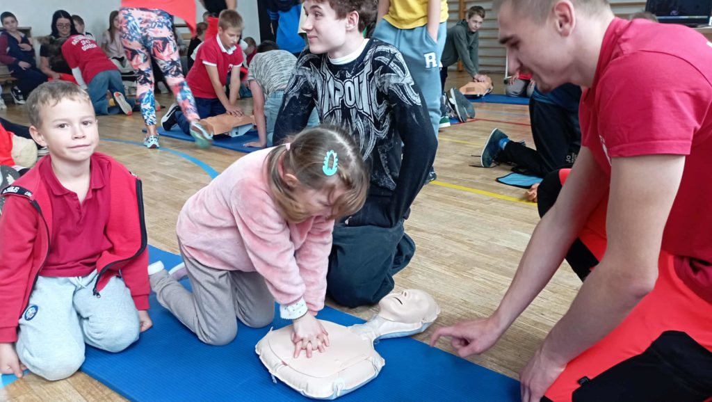 Na pierwszym planie uczennica pierwszej klasy uciskająca klatkę fantoma obok jej kolega z klasy i uczeń 8 klasy. Przed nimi ratownik, za nimi inni ćwiczący RKO.