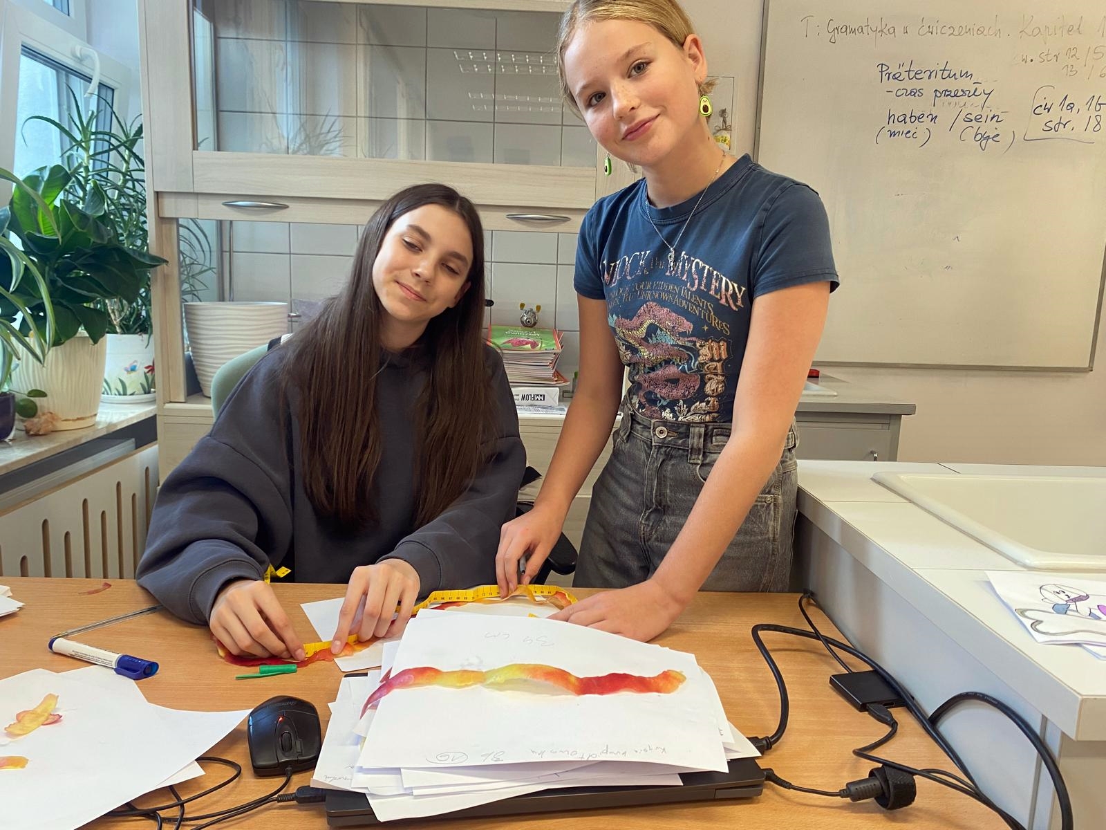 Dwie dziewczynki: po lewej siedzi za biurkiem, po prawej stoi obok. Obie trzymają miarkę i mierzą obierkę jabłka leżącą na stosie papierów przed nimi.