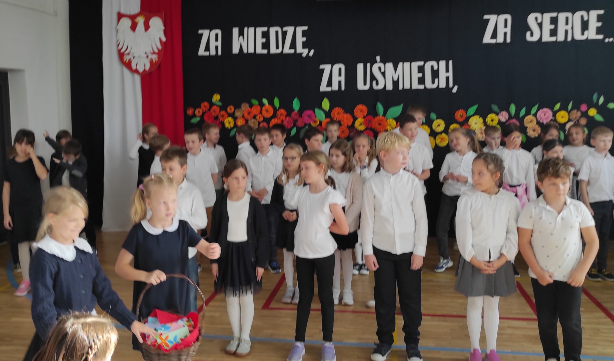 Na pierwszym planie stoją dwie dziewczynki, jedna z nich trzyma koszyczek w dłoni. Na drugim planie stoi 6, ustawionych w rzędzie uczniów.  Za nimi stoją  ustawione w trzech rzędach dzieci. Wszyscy są ubrani w stroje galowe. W tle dekoracja: zawieszona pionowo flaga Polski, na niej godło,  dalej napis i kwiaty.