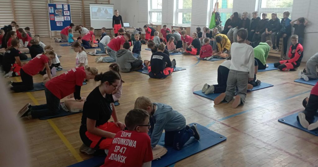 W sali gimnastycznej na podłodze leżą 3 rzędy fantomów przy nich klęczą po 2 lub 3 osoby. Jedna z osób wykonuje na fantomie masaż serca. W tle stolik z laptopem i rzutnikiem oraz tablica z plakatem RKO.