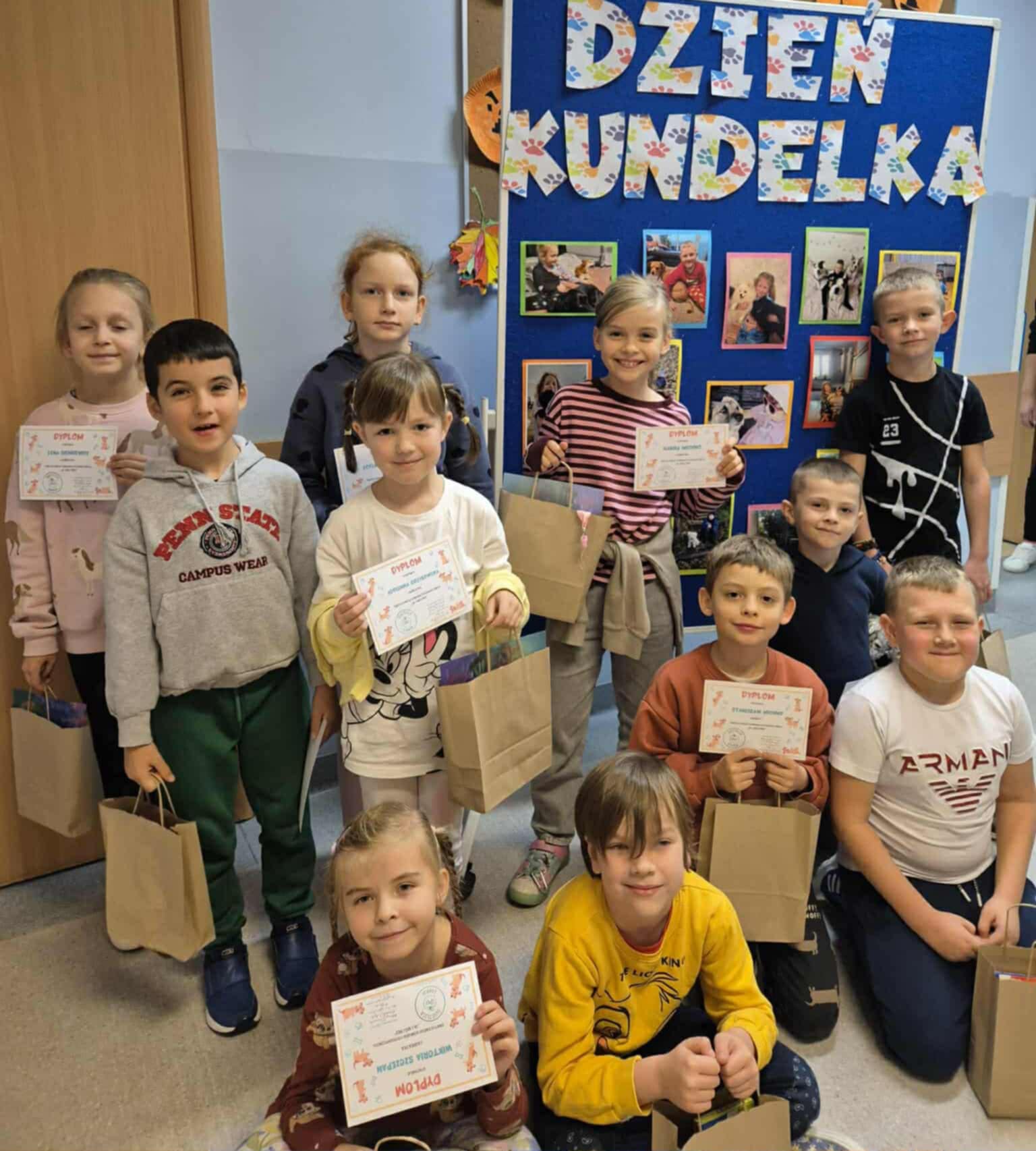 Grupa uczniów pozuje przed tablicą z napisem dzień kundelka „Dzień Kundelka”. Dzieci trzymają dyplomy i papierowe torebki z nagrodami.