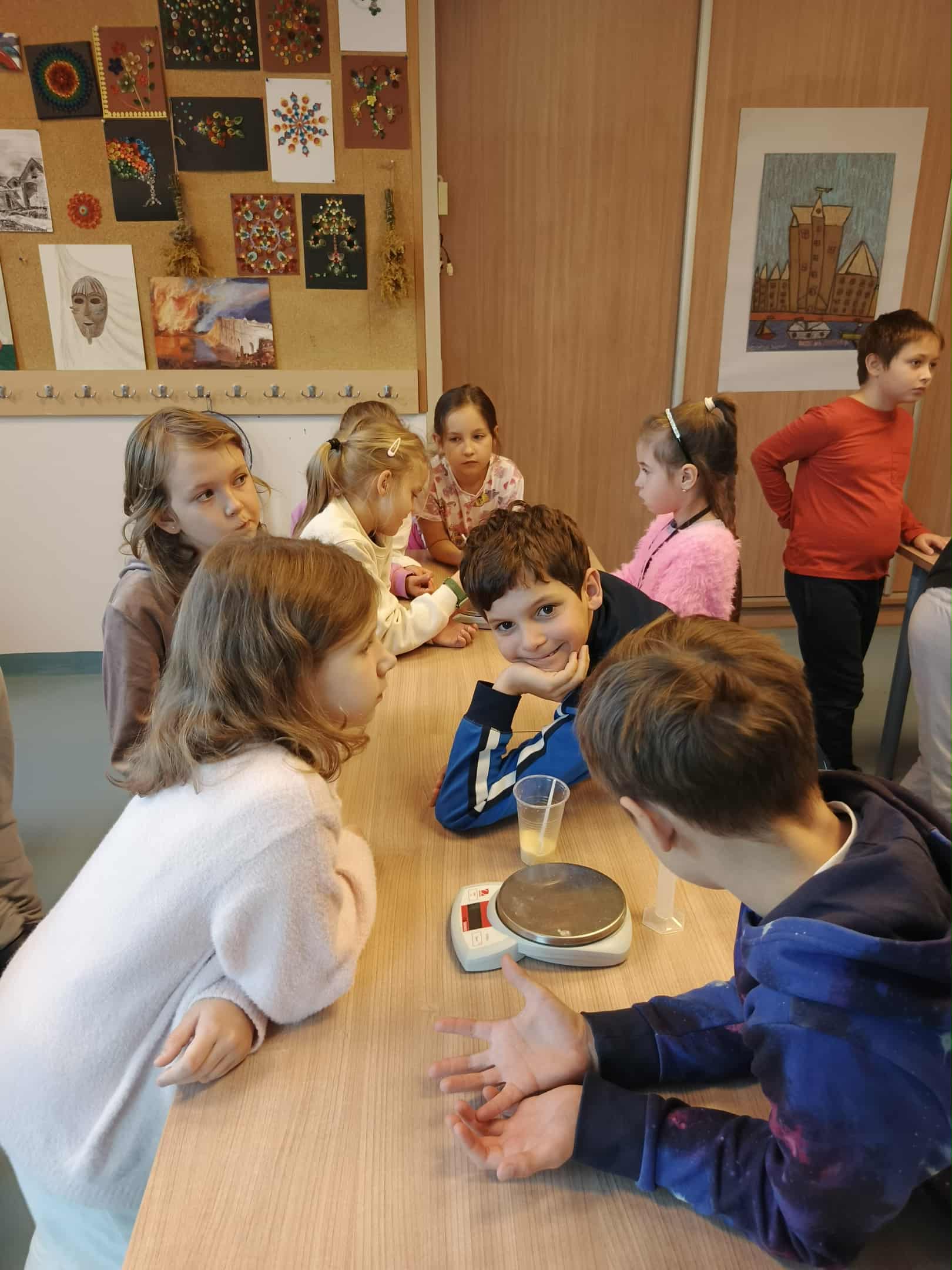 W sali stoi kilkoro dzieci przy stole. Na stole leży waga elektroniczna, przezroczysty kubek i mała miarka. W tle widać tablicę z kolorowymi pracami plastycznymi.
