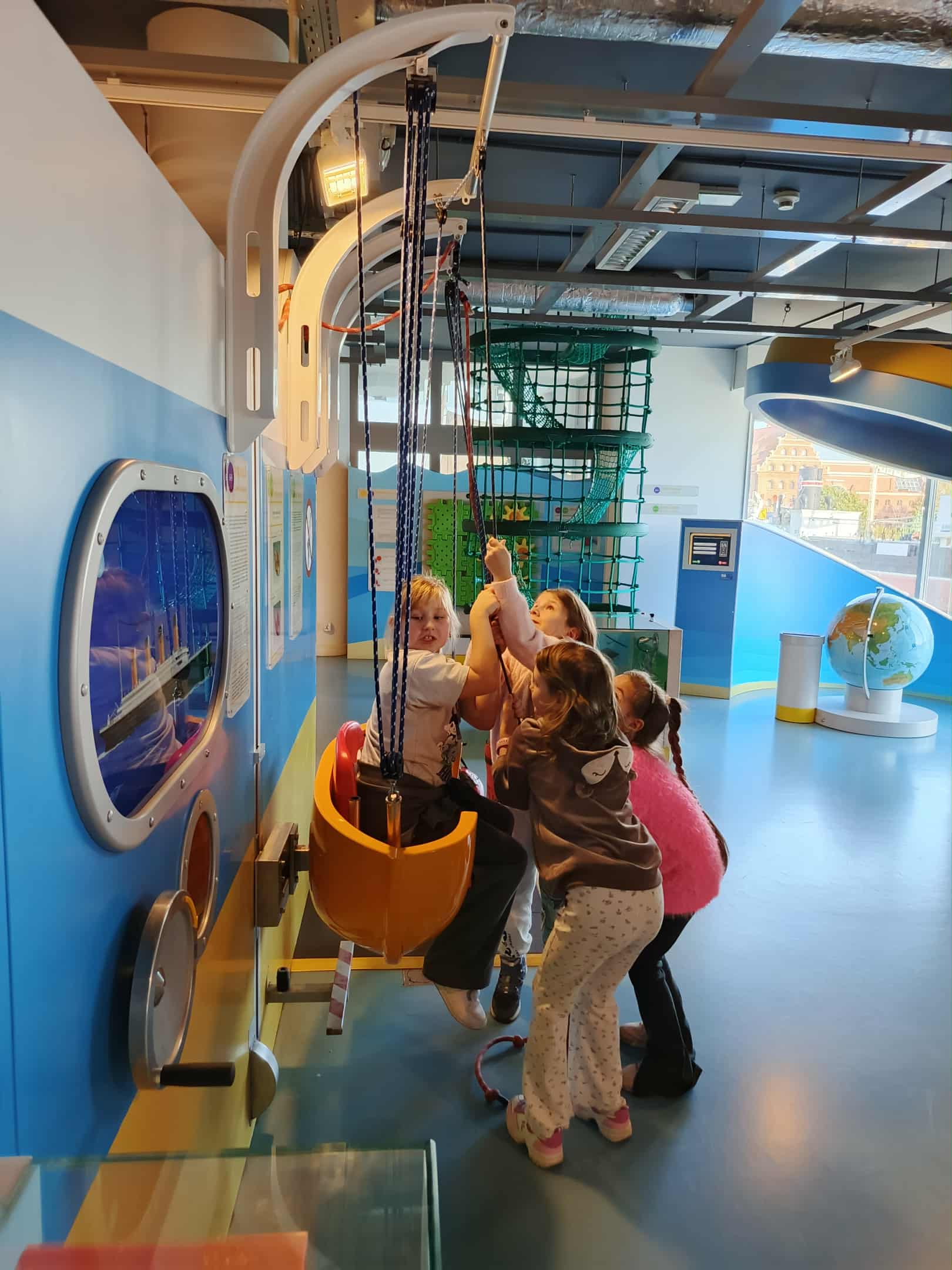 Cztery dziewczynki bawią się przy stanowisku z linami i fotelem zawieszonym na hakach. Jedna dziewczynka siedzi w fotelu, a trzy inne ciągną za liny, żeby ją unieść do góry. W tle widać kolorowe ściany z niebieskimi i żółtymi elementami oraz wystawy związane z morzem.